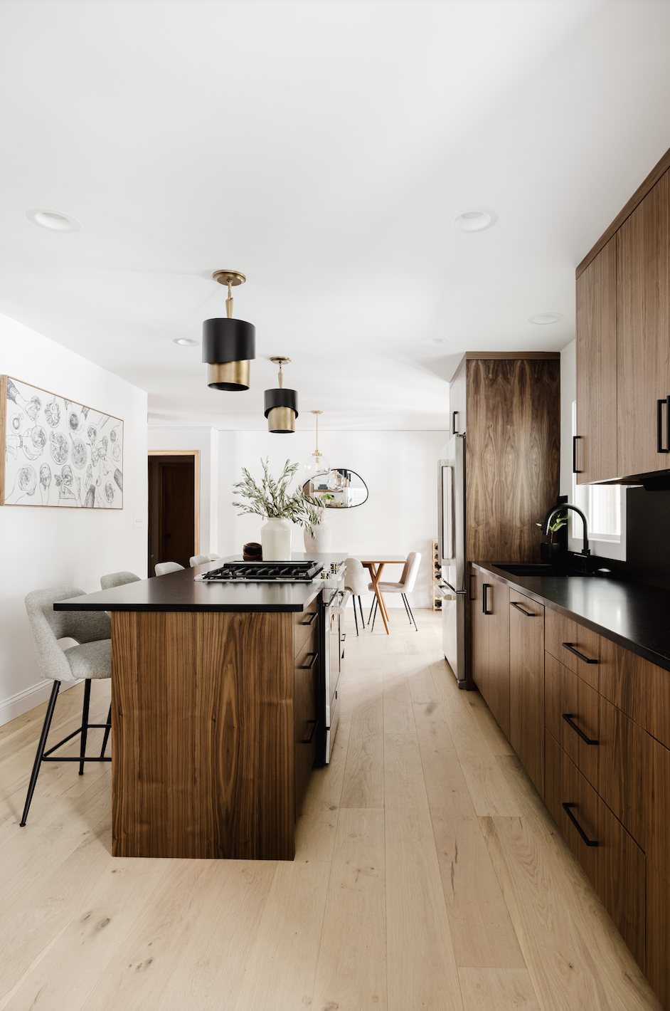 Wide view of custom walnut kitchen cabinetry featuring integrated appliance panels, a black undermount sink, and a long wood-paneled island with a built-in gas range.
