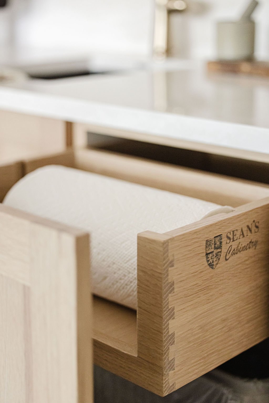 High-angle close-up of a custom light oak pull-out drawer featuring a paper towel roll holder. The side of the drawer showcases precise dovetail joinery and a laser-engraved "Sean's Cabinetry" logo, with a white quartz countertop and gold faucet visi