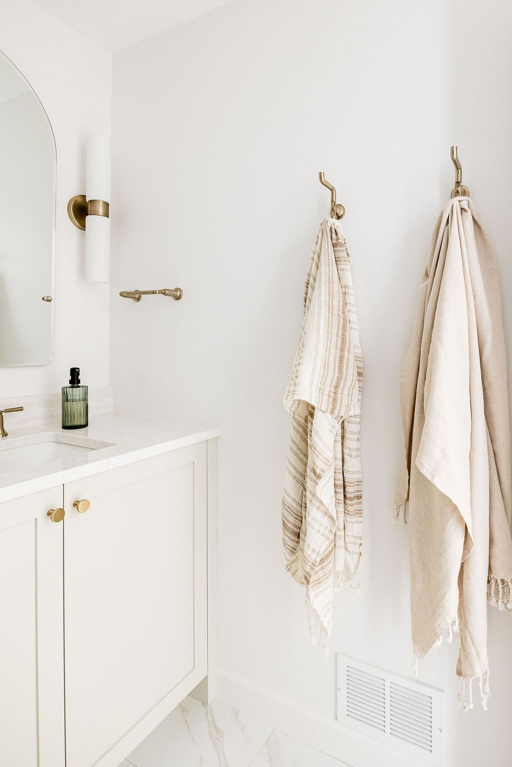 Bright bathroom featuring a white shaker vanity with gold knobs and a white marble countertop. Two brass wall hooks hang to the right of the vanity, holding neutral-toned fringed towels against a clean white wall.