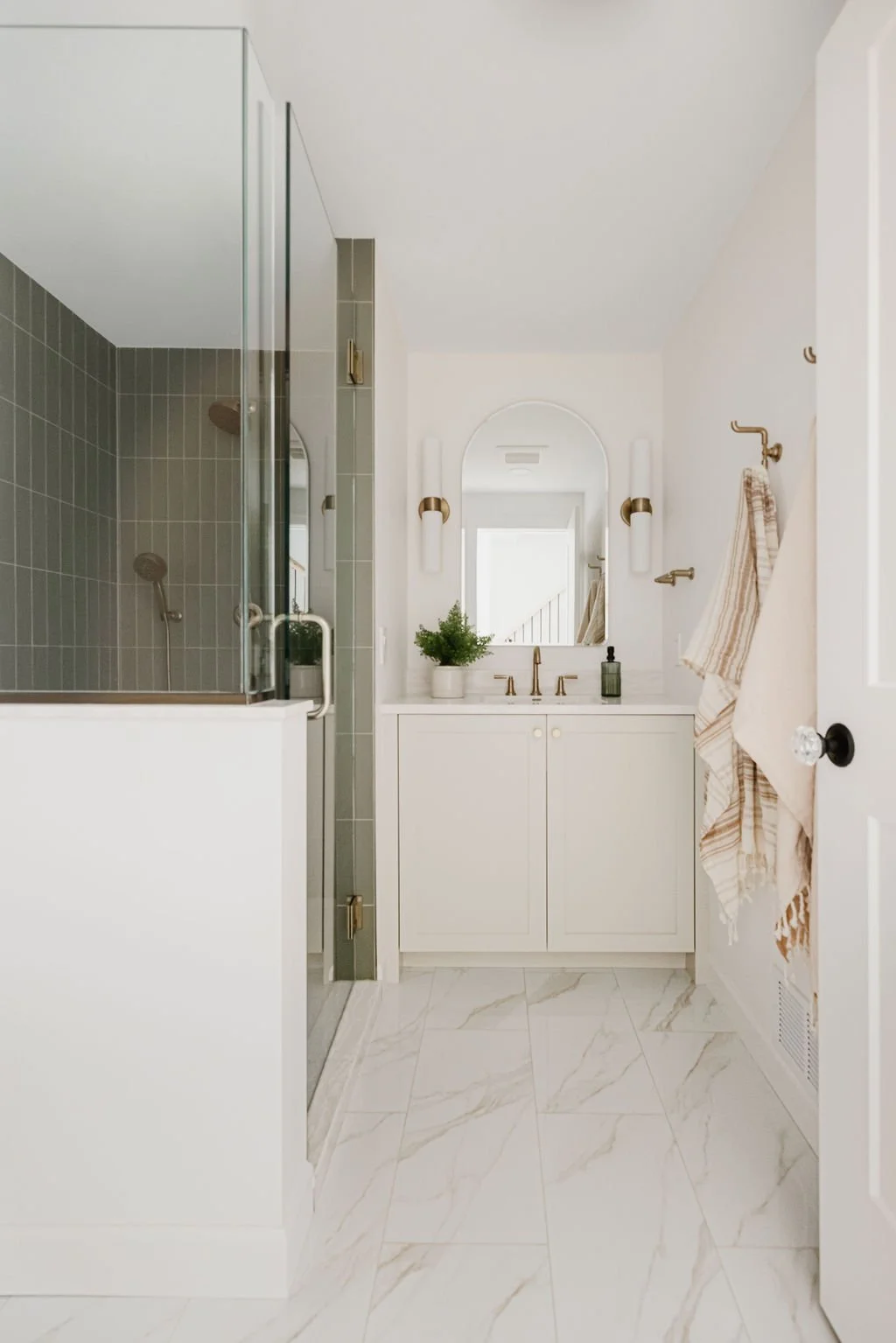 A wide view of a modern bathroom featuring marble floor tiles and a custom white shaker vanity with gold hardware. The layout includes a glass walk-in shower with vertical green tiling and neutral-toned towels hanging on brass wall hooks.