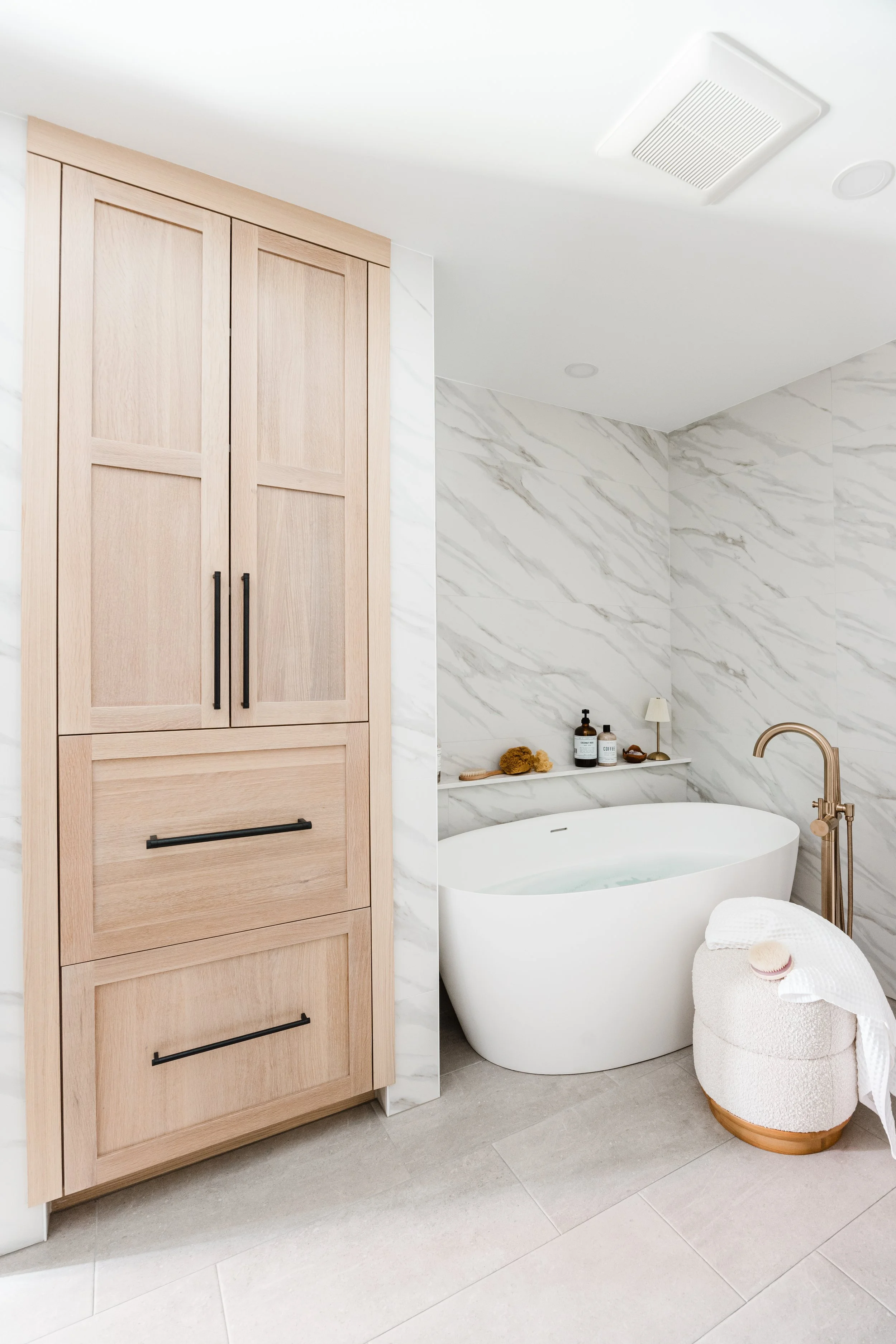 Light wood cabinets with black hardware next to a freestanding bathtub and marble tile