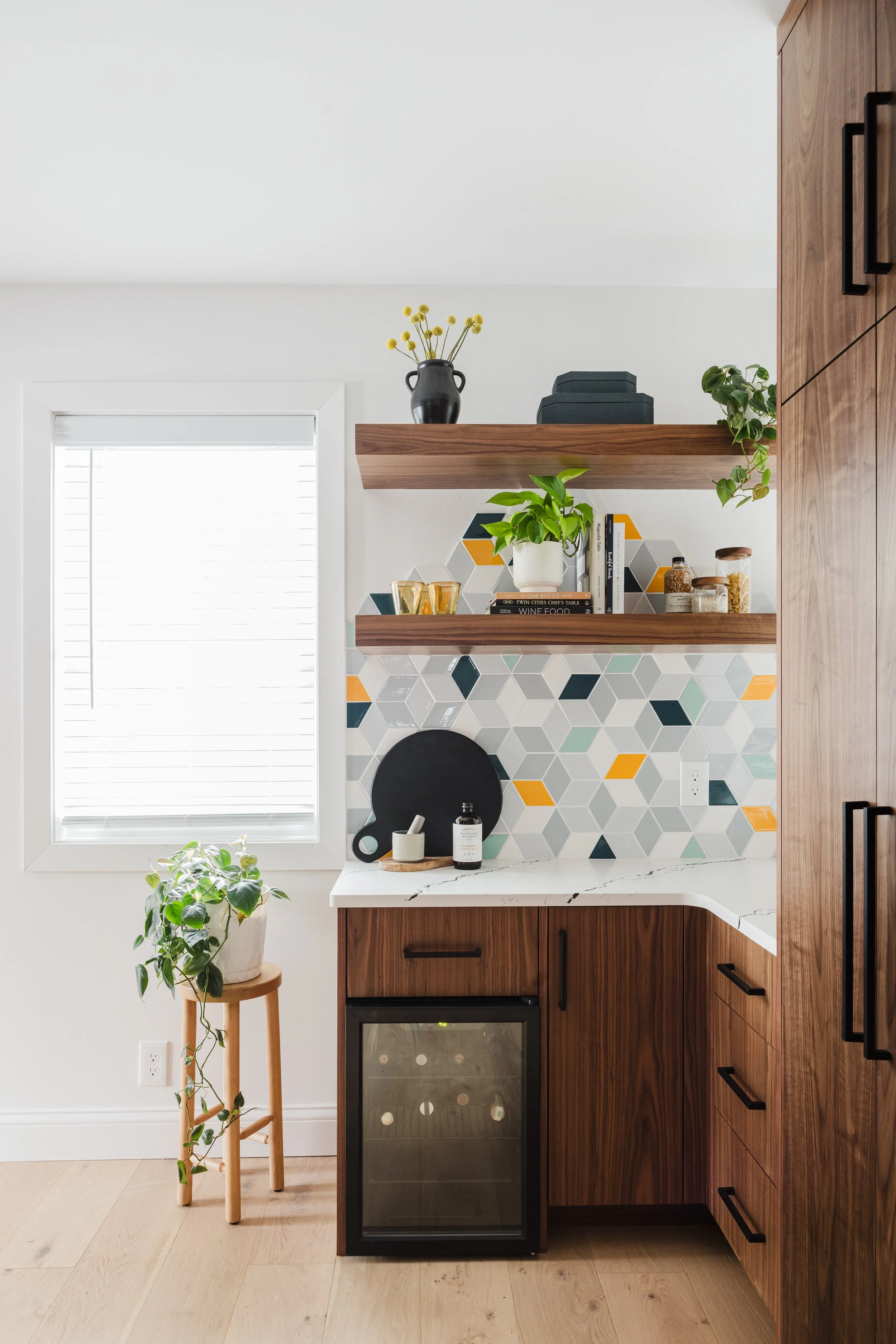 Close-up of a custom kitchen coffee station featuring a geometric cube-patterned backsplash in gray, blue, and yellow. The space includes a built-in wine fridge, walnut-toned base cabinets with black pulls, white quartz countertops, and two matching 