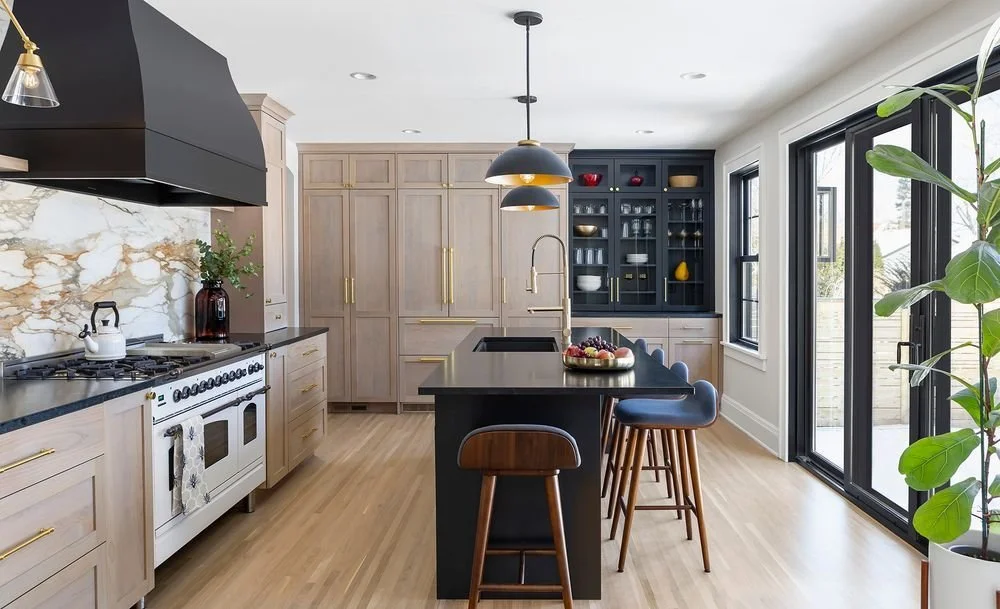 Modern kitchen with light wood shaker cabinets, a matte black island with waterfall edge, brass hardware, and floor-to-ceiling pantry storage, accented by large black-framed windows and glass-front navy built-ins.