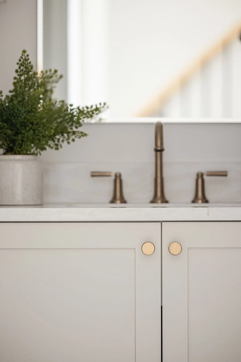 Close-up of light gray shaker-style bathroom cabinets featuring round brushed gold knobs, a white quartz countertop, and a matching gold faucet.