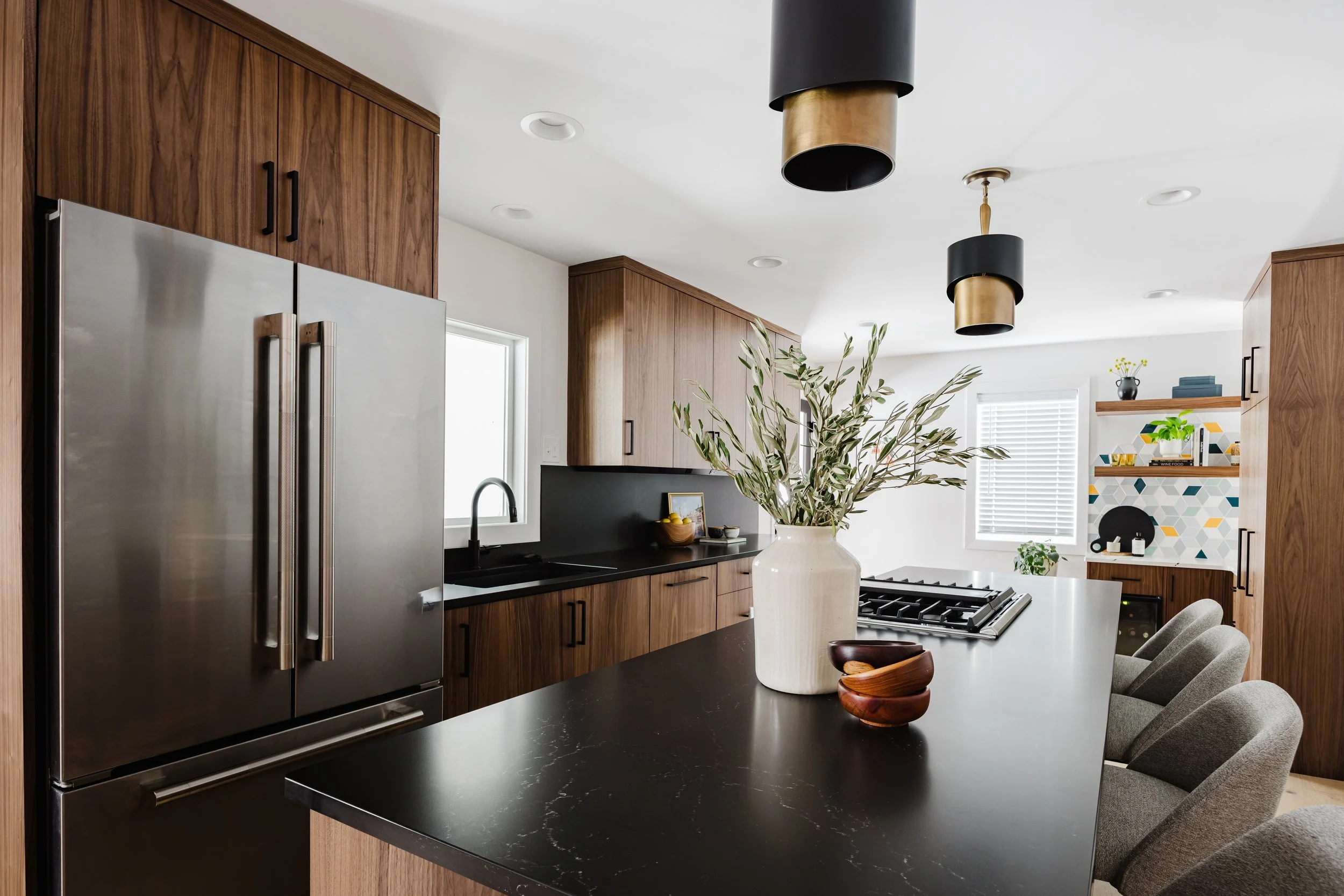 Wide perspective of a custom wood kitchen island with a dark quartz countertop and integrated gas range. In the background, a built-in coffee station features geometric-patterned tile, floating wood shelves, and matching walnut-toned cabinetry.