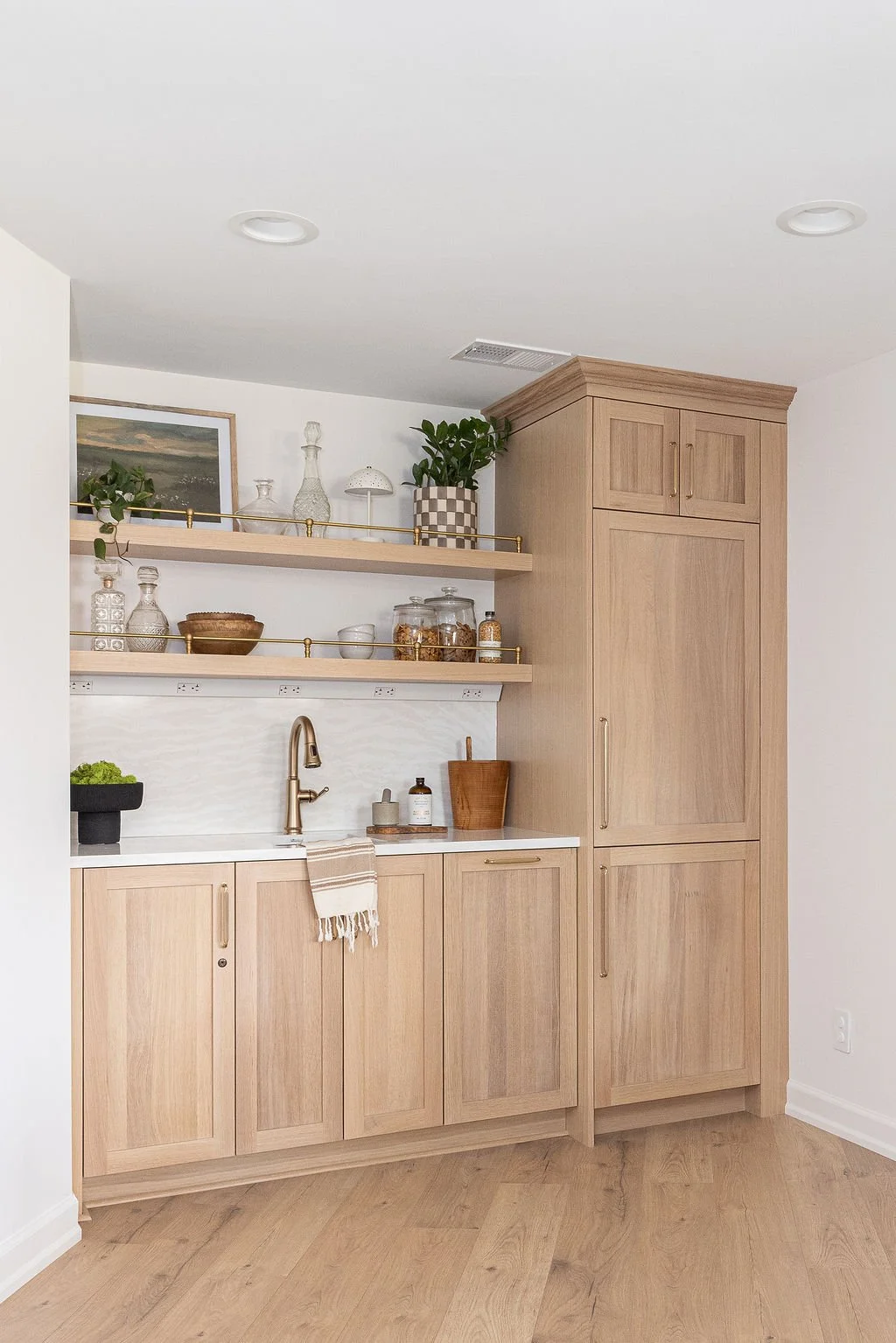 Custom light oak wet bar featuring flat-panel cabinets and a tall integrated refrigerator unit with crown molding. Two floating oak shelves with brass gallery rails display decor above a white marble countertop and gold faucet.