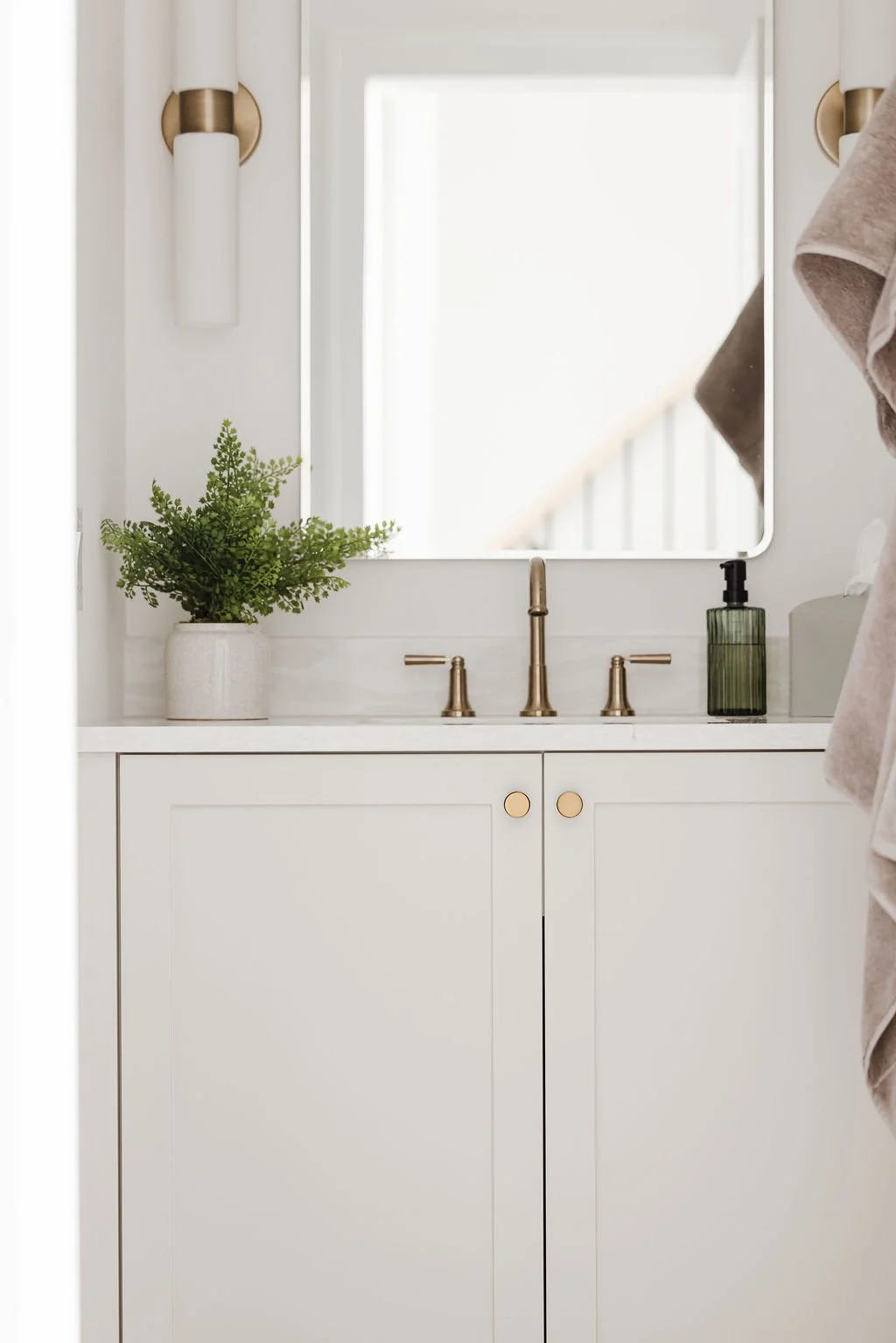 Front-facing view of a white shaker-style vanity featuring circular brass knobs and a matching gold faucet set. Symmetrical white tube sconces with brass accents frame a rectangular mirror above the clean white countertop.