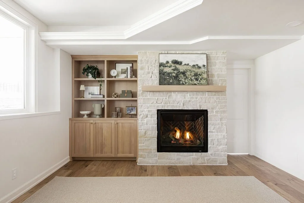 Custom light wood built-in cabinet with open shelving beside a white stone fireplace with wood mantel in a bright, modern living room