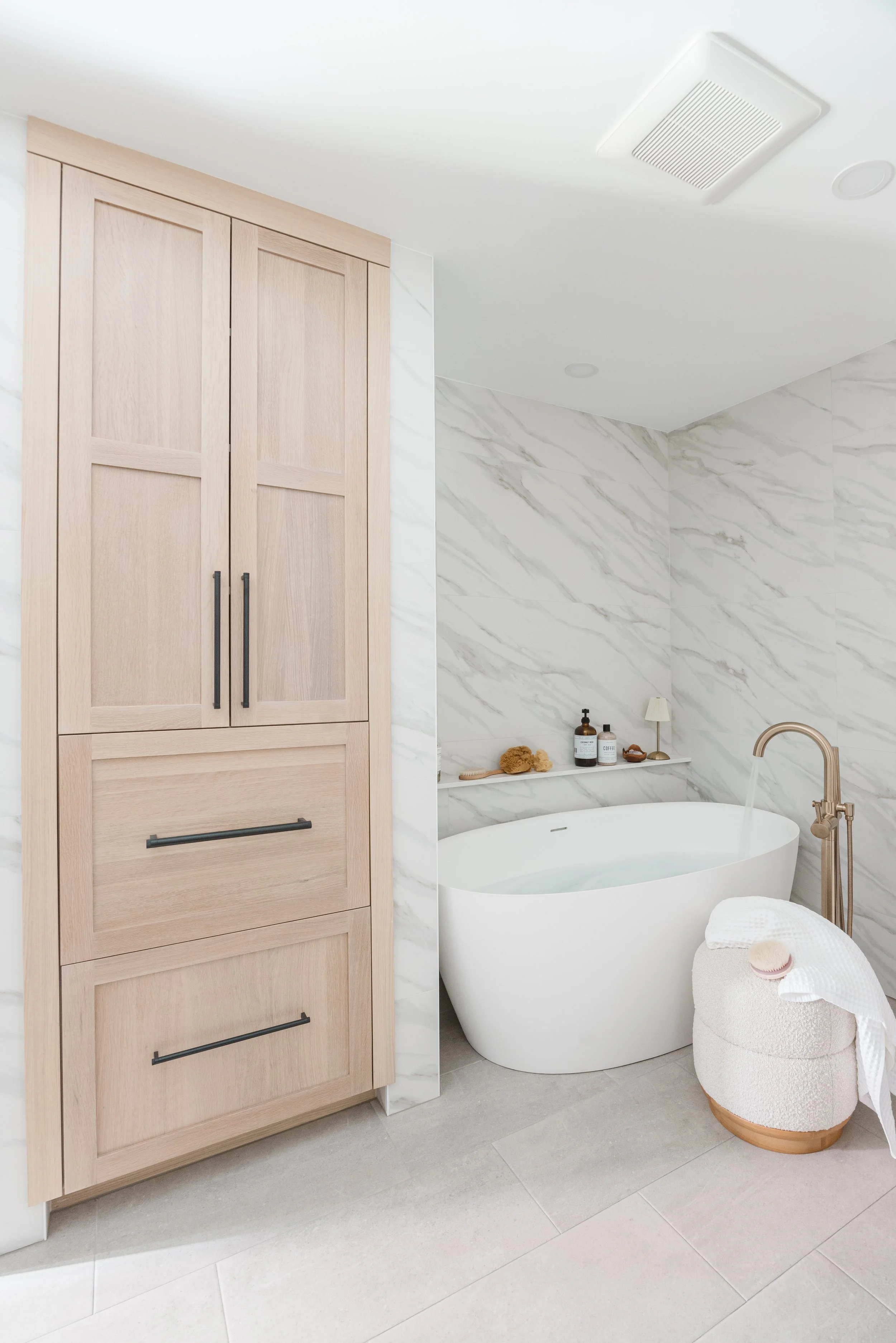 Light wood bathroom cabinets beside a freestanding tub and marble walls