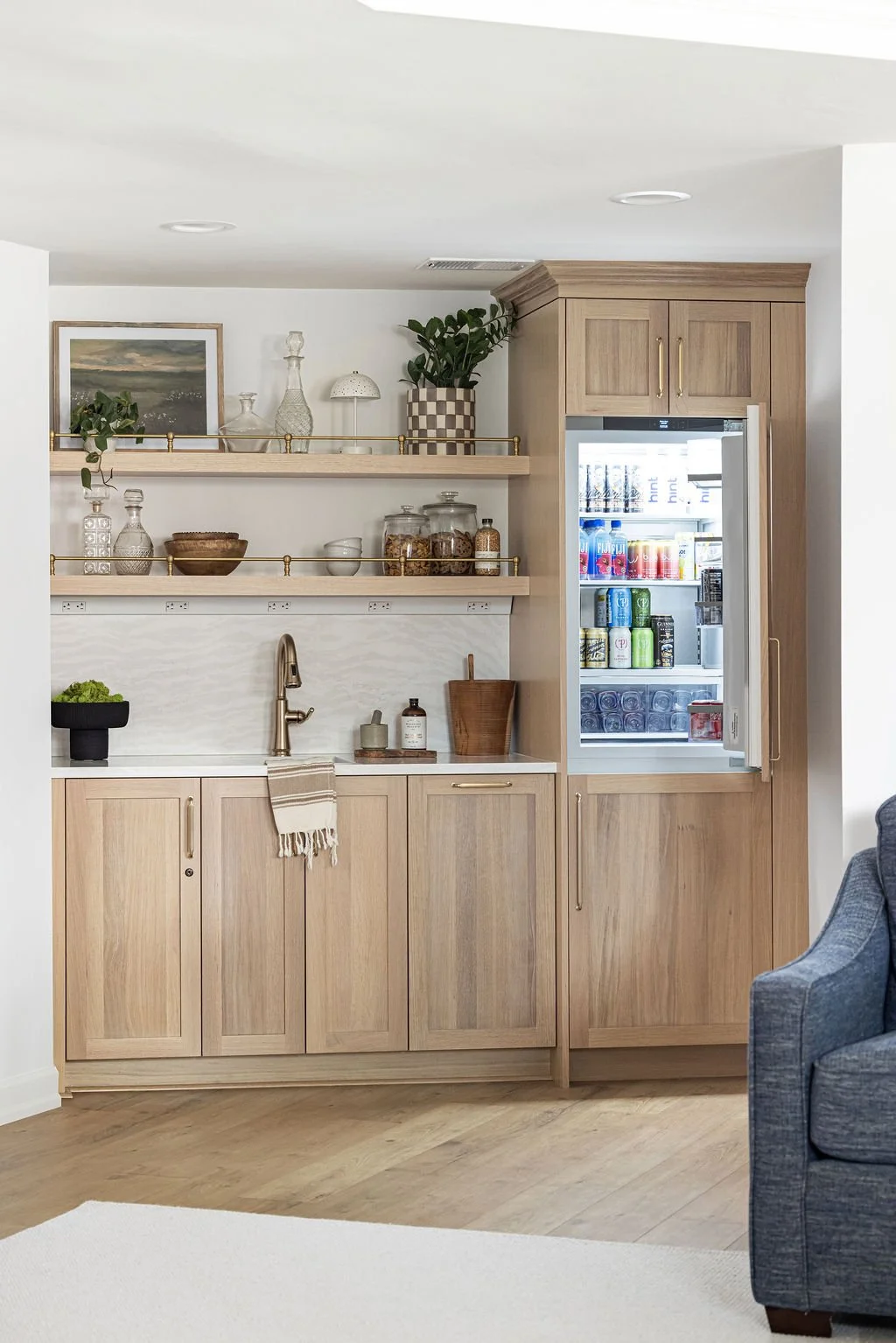 Custom light oak wet bar featuring flat-panel lower cabinets and an open integrated refrigerator column. Two matching floating shelves with gold gallery rails display glassware and decor against a white tiled backsplash.