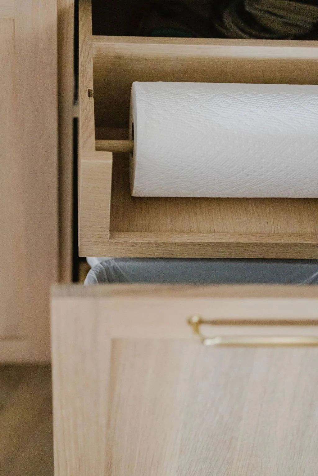 Detailed vertical shot of a custom light oak pull-out cabinet. The top section features an integrated wooden paper towel holder with a visible dowel, while the bottom section reveals a pull-out waste bin. The flat-panel cabinetry is finished with a s