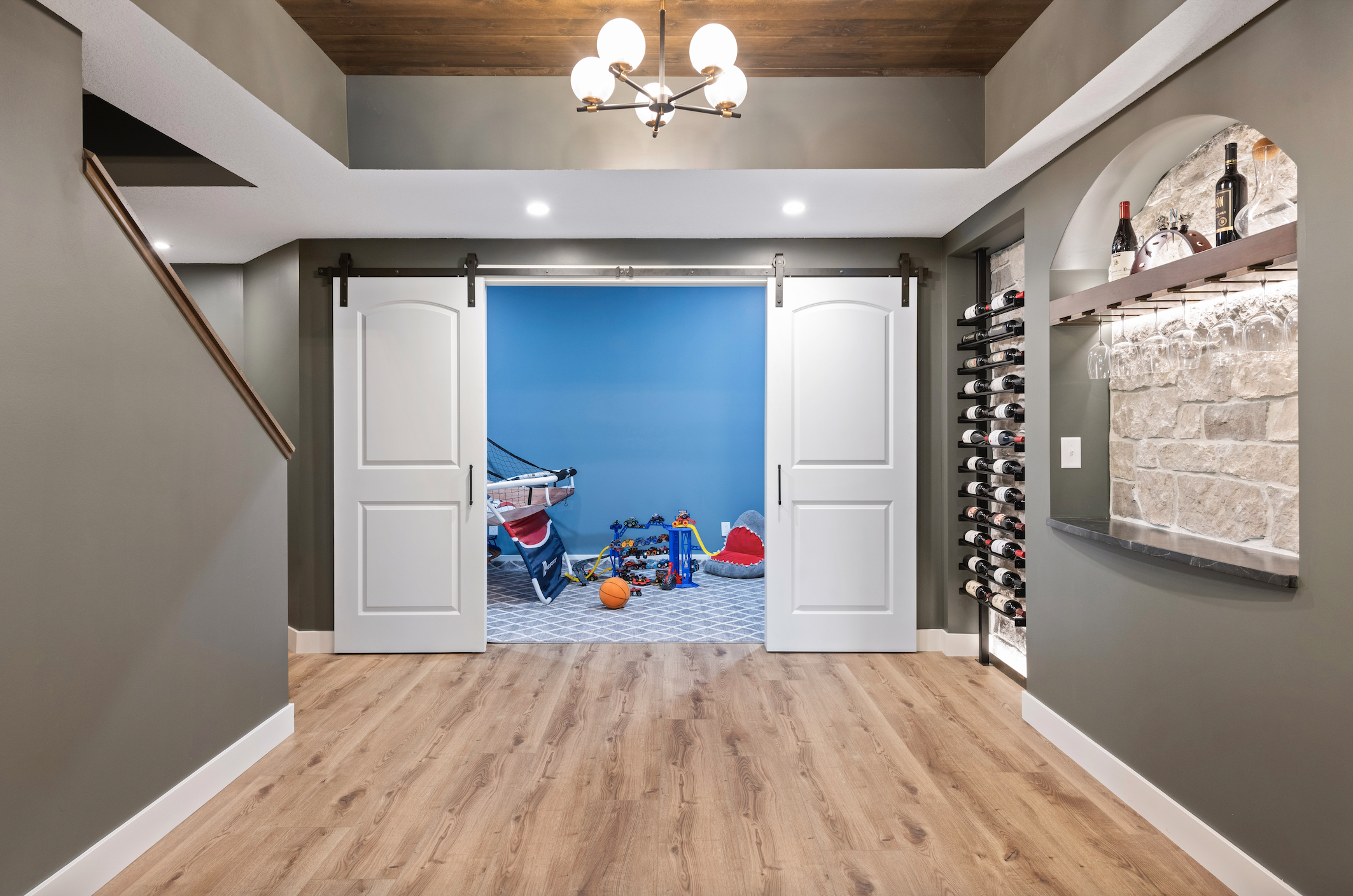Custom white sliding barn doors on black hardware open to a children’s playroom, flanked by built-in wine storage and stone display shelving.