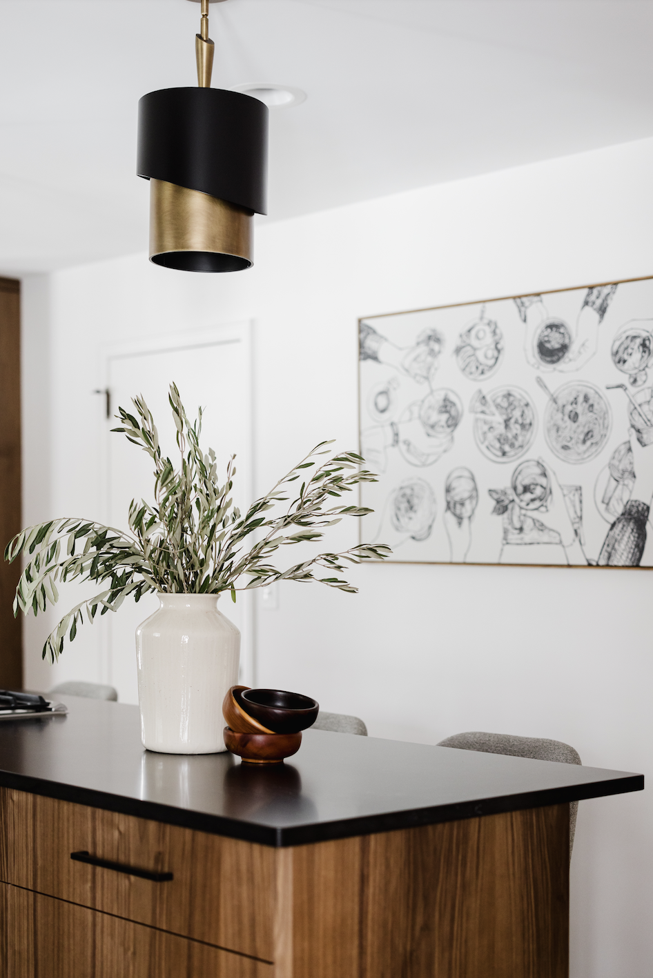 Custom walnut kitchen island with dark stone countertops, featuring a white decorative vase and modern black and gold pendant lighting against a minimalist white wall.
