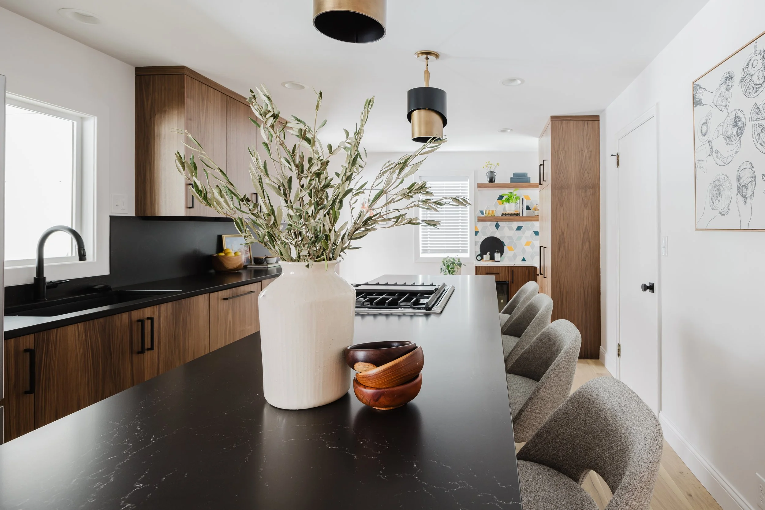 Wide-angle view of a modern kitchen featuring a dark stone-top island with seating and custom wood-slab cabinetry. The background shows a decorative tiled coffee station with open wood shelving and integrated appliance panels.
