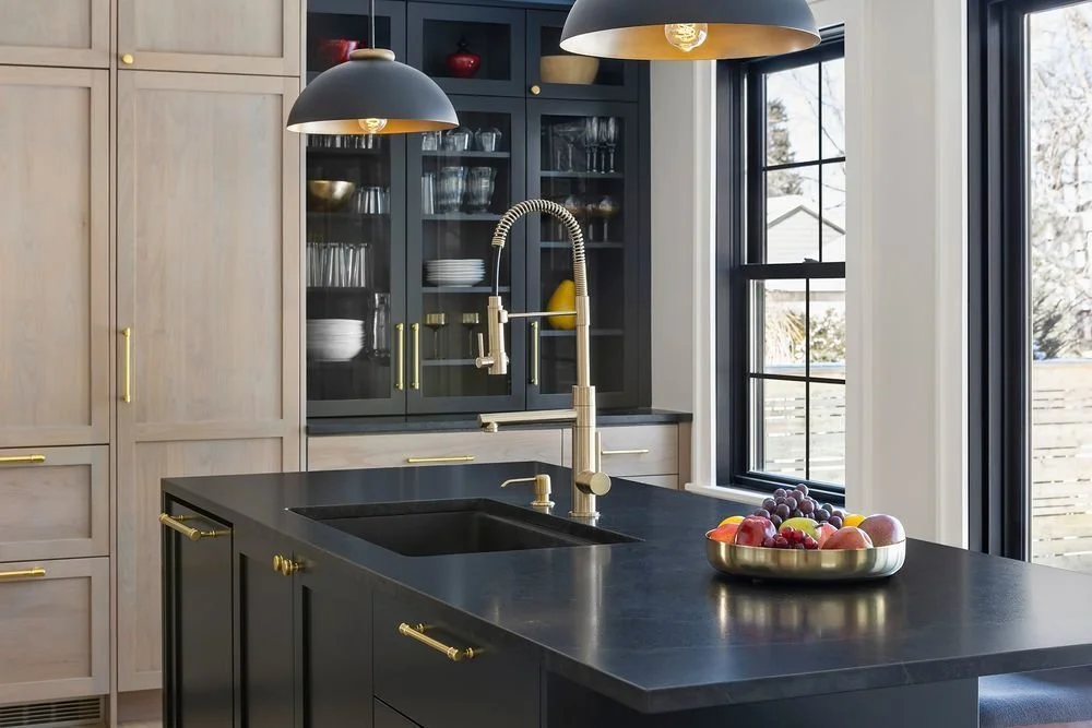 Dark brown and black kitchen island with brass fixtures, integrated sink, and glass-front upper cabinets in deep charcoal finish.