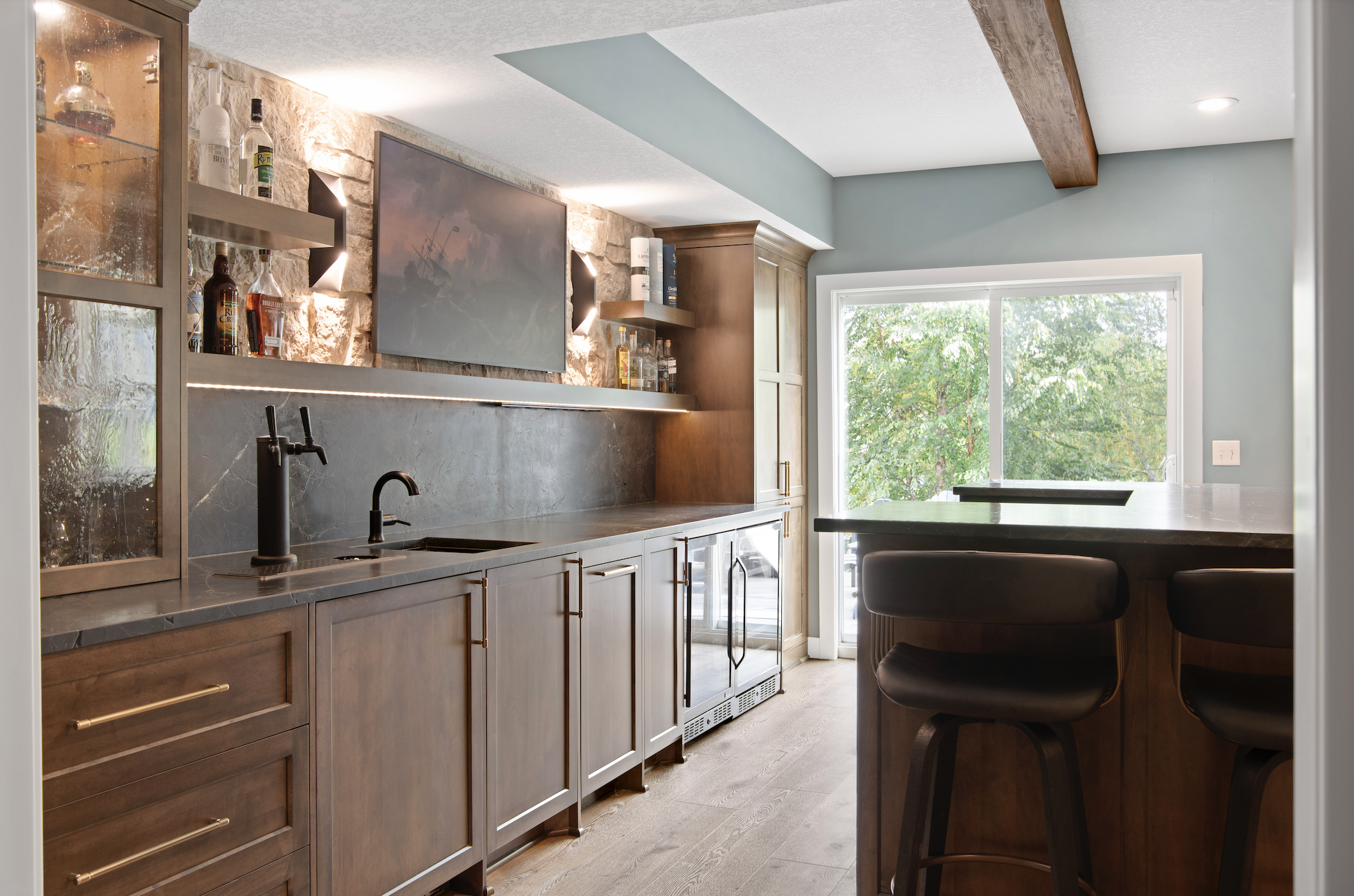 Custom home bar with dark wood cabinetry, stone backsplash, open shelving with accent lighting, matte black faucet, and a waterfall island with bar seating and large window.