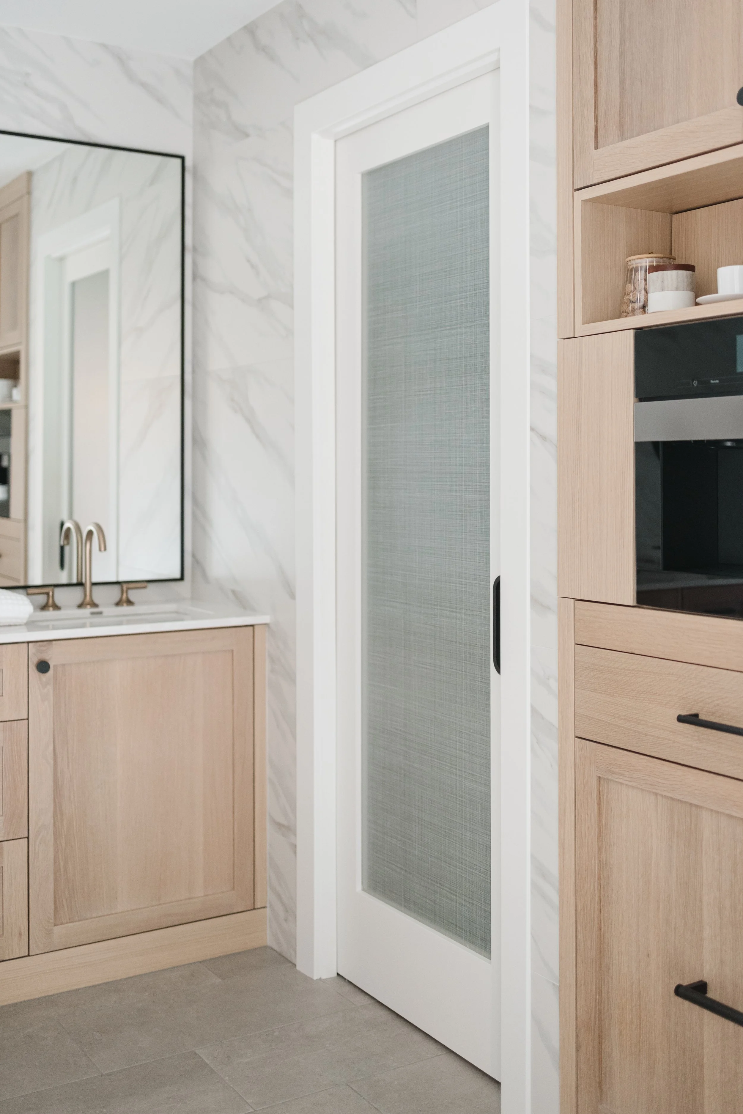 Light wood cabinets with sleek hardware beside a frosted glass door and marble backsplash in a modern space