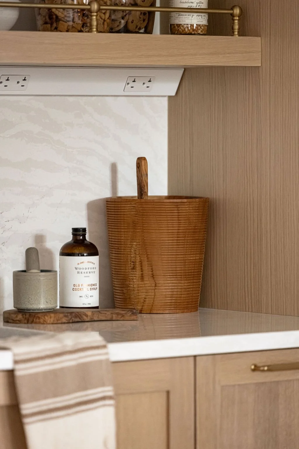 A close-up detail of a light oak wet bar featuring a fluted wooden ice bucket and cocktail ingredients on a white quartz countertop. It's framed by flat-panel oak cabinetry and a white marble-patterned backsplash under a floating shelf with a brass g