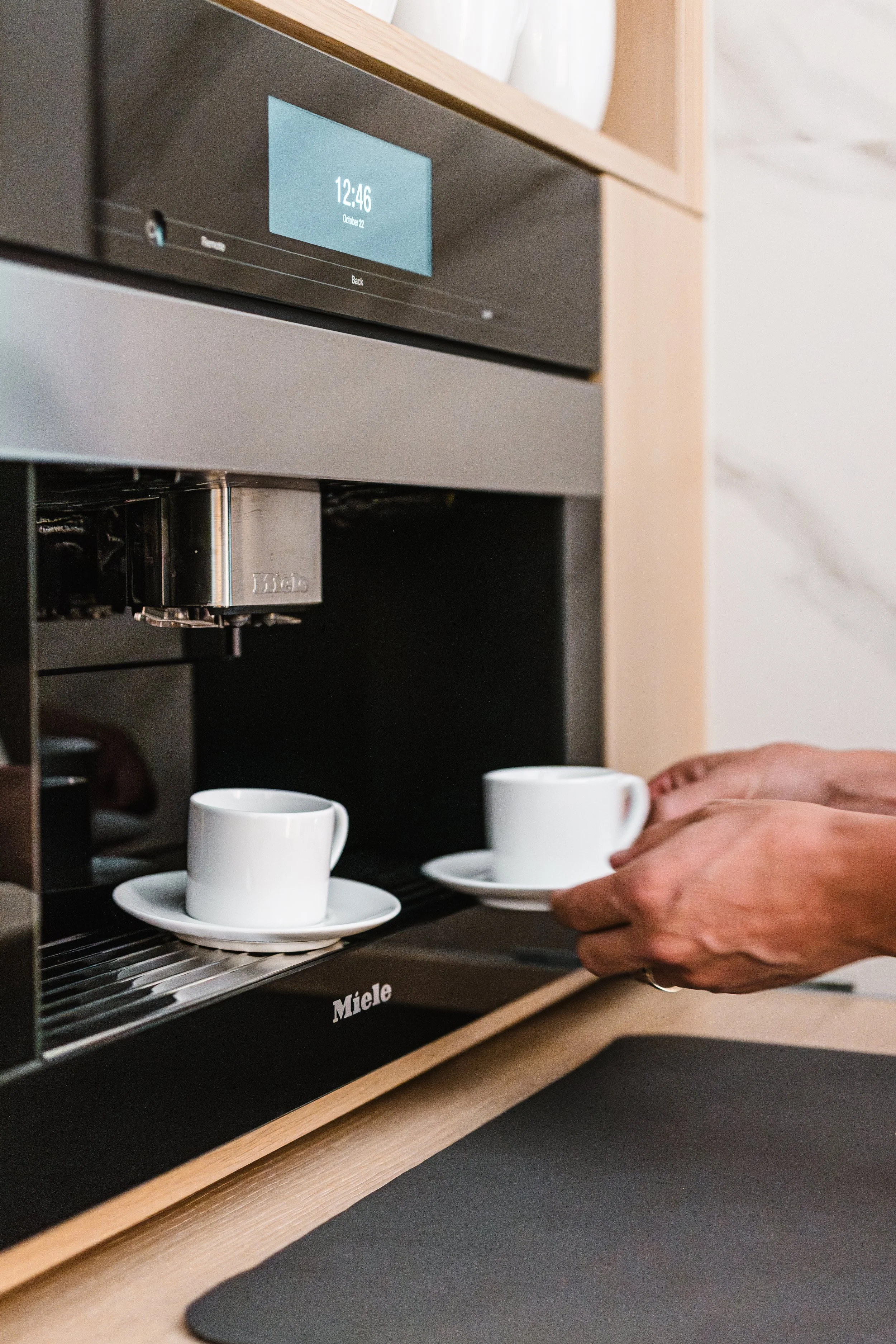 Built-in coffee machine set within light wood cabinets with two cups being placed