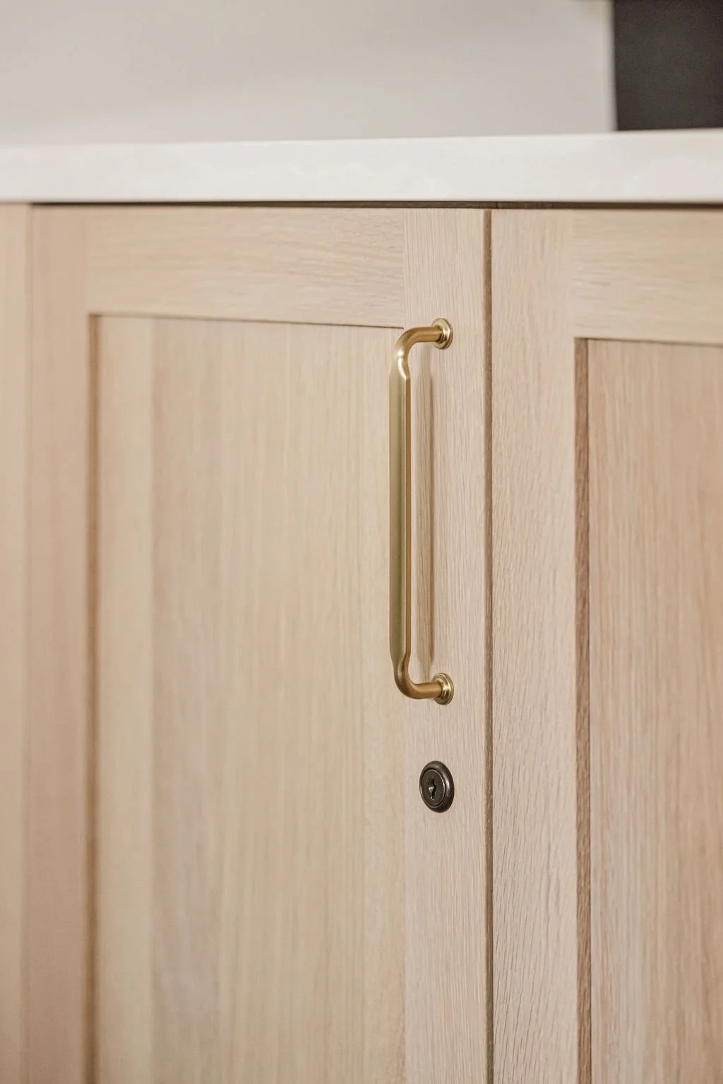 Close-up of custom light oak cabinetry featuring a slim, modern brass pull handle. The flat-panel doors showcase a natural wood grain and include a small integrated lock detail below the hardware.