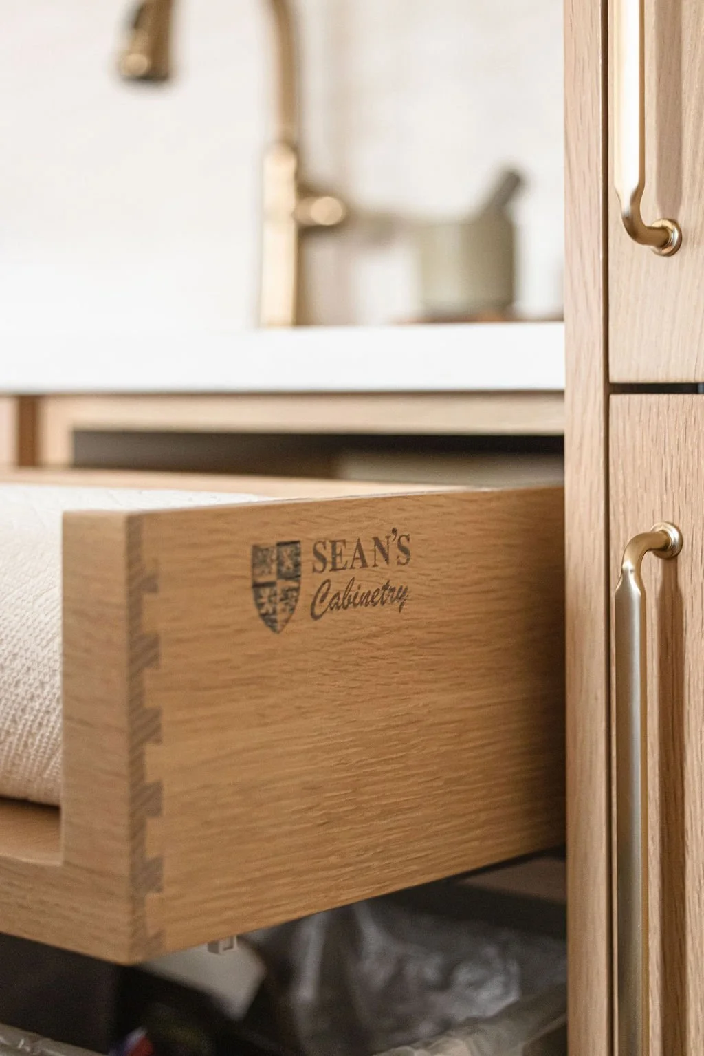 Detailed close-up of a custom light oak pull-out drawer featuring a laser-engraved "Sean's Cabinetry" logo on the side. The drawer is built with high-quality dovetail joinery and is shown partially open with a white paper towel roll inside.