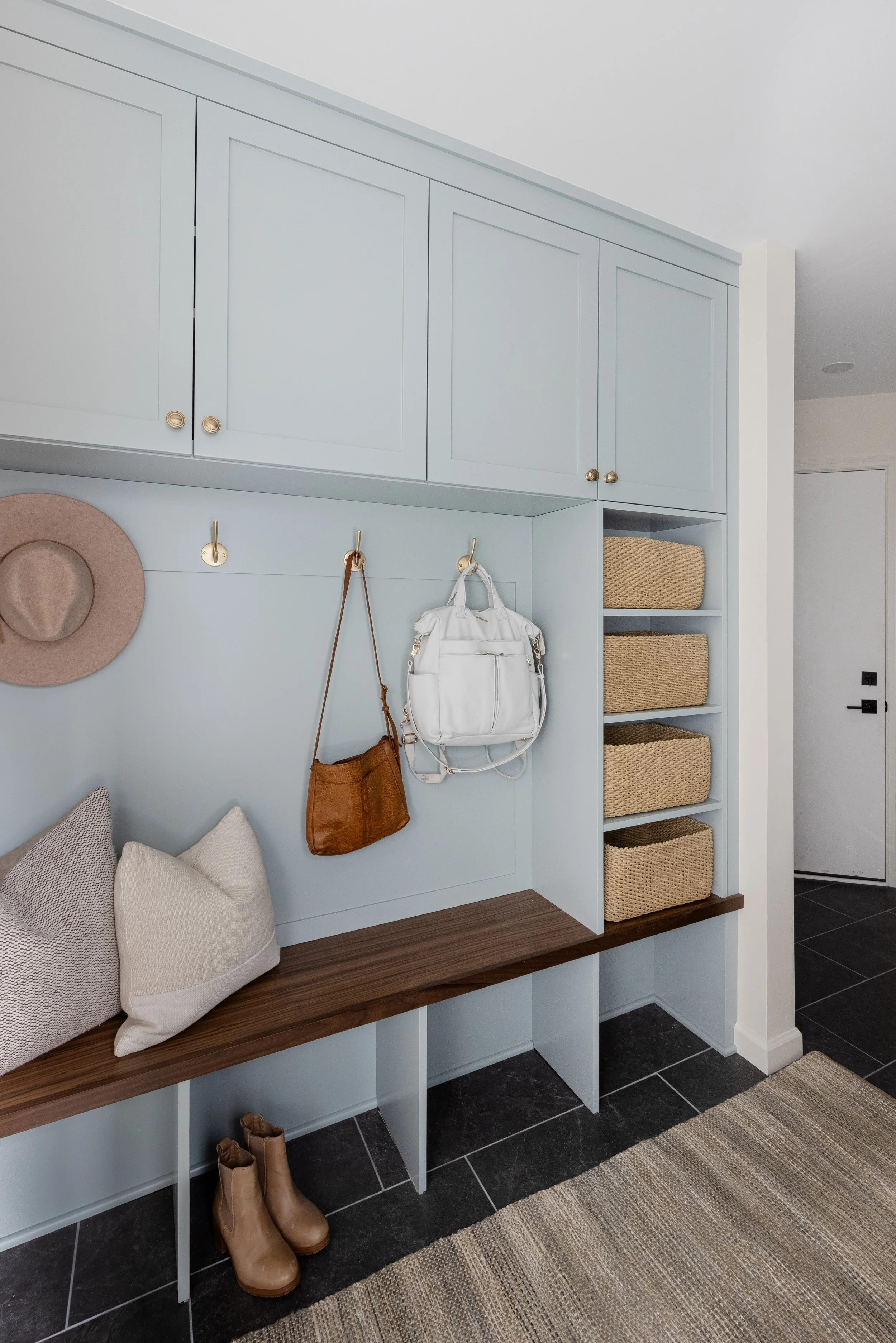 Custom mudroom built-ins with light blue shaker cabinets, wood bench, brass hooks, and woven basket storage