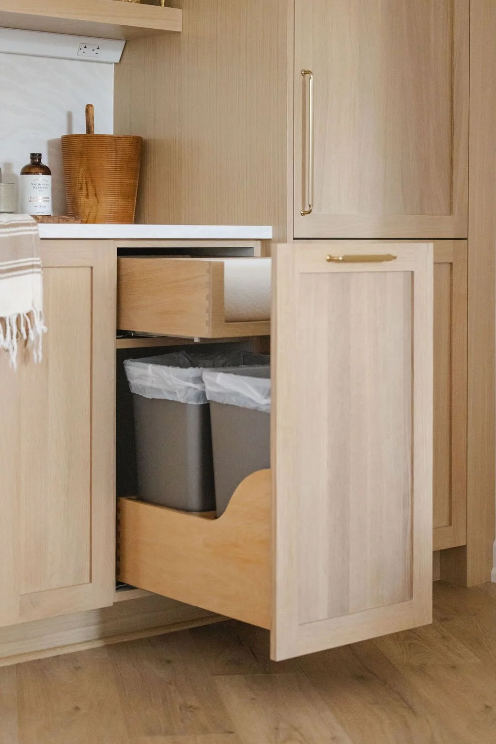 Perspective view of an open custom light oak cabinet featuring a dual-tiered pull-out system. The top tier holds an integrated paper towel roll, while the bottom tier houses a double trash and recycling bin unit. The shaker-style cabinet door is equi