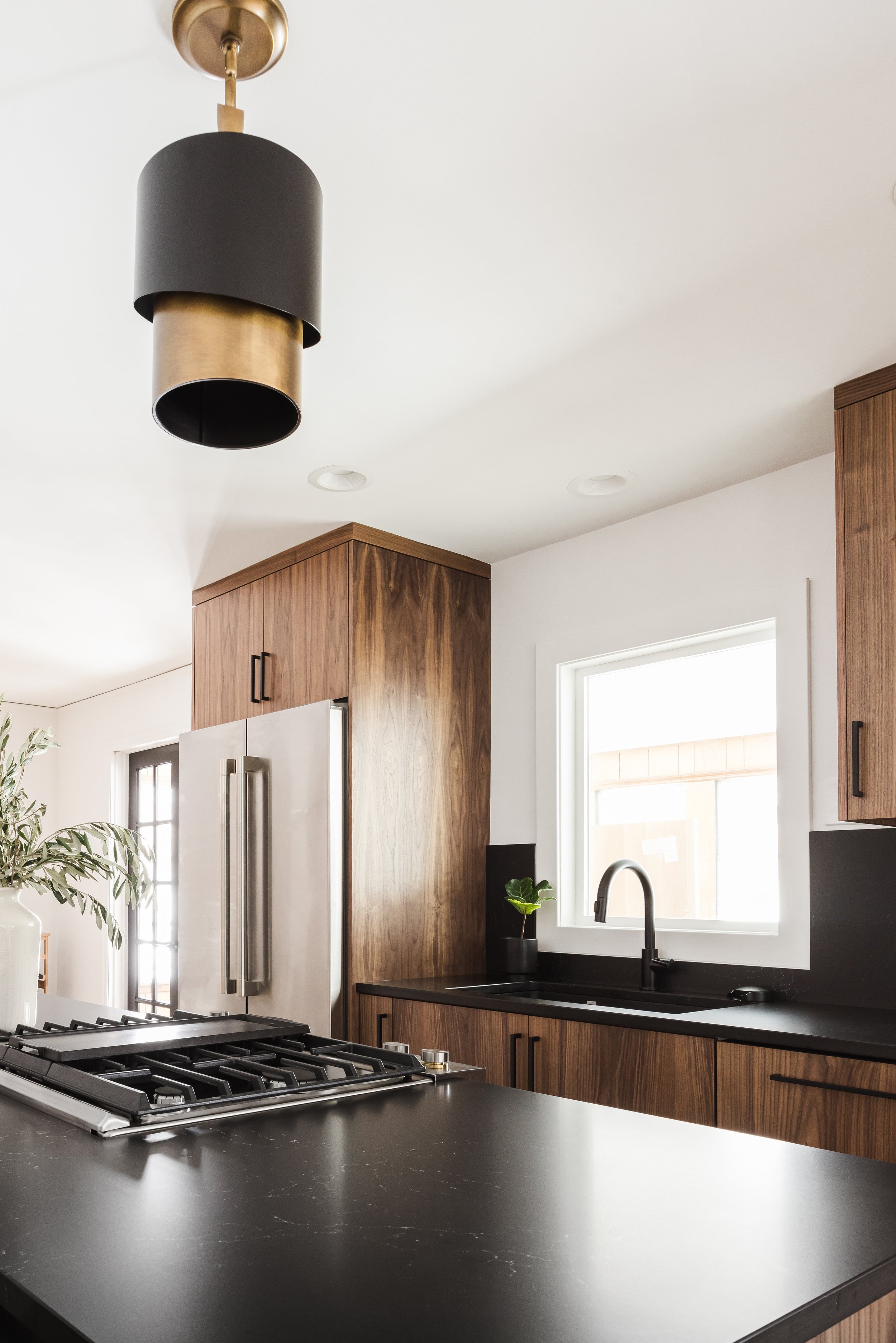 Close-up of a kitchen island with a dark countertop, focusing on the custom wood cabinetry and integrated range. The view looks toward a window and a black sink, highlighting the interplay between natural wood grain and matte black fixtures.