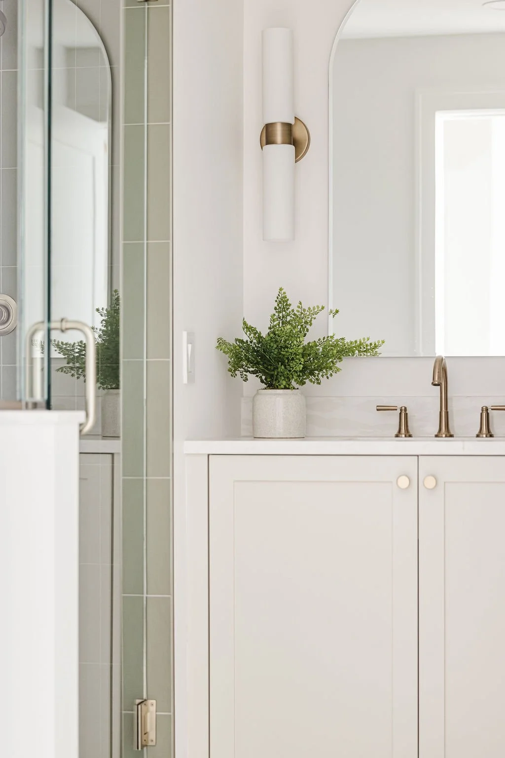 Custom white shaker-style bathroom vanity with round brass knobs, an arched mirror, and white quartz countertops paired with a glass walk-in shower.
