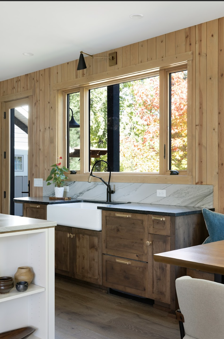 Custom wood kitchen base cabinets with farmhouse sink, marble backsplash, and black countertop beneath a large window.