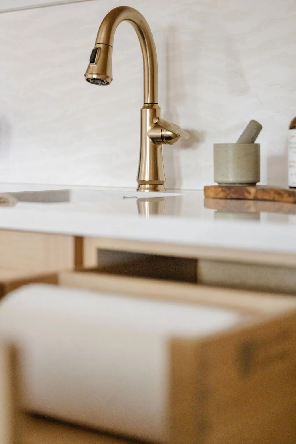 Close-up of a light oak wet bar with a white countertop featuring an integrated black undermount sink. An open pull-out drawer specifically designed for a paper towel roll reveals the functional custom cabinetry below a gold gooseneck faucet.