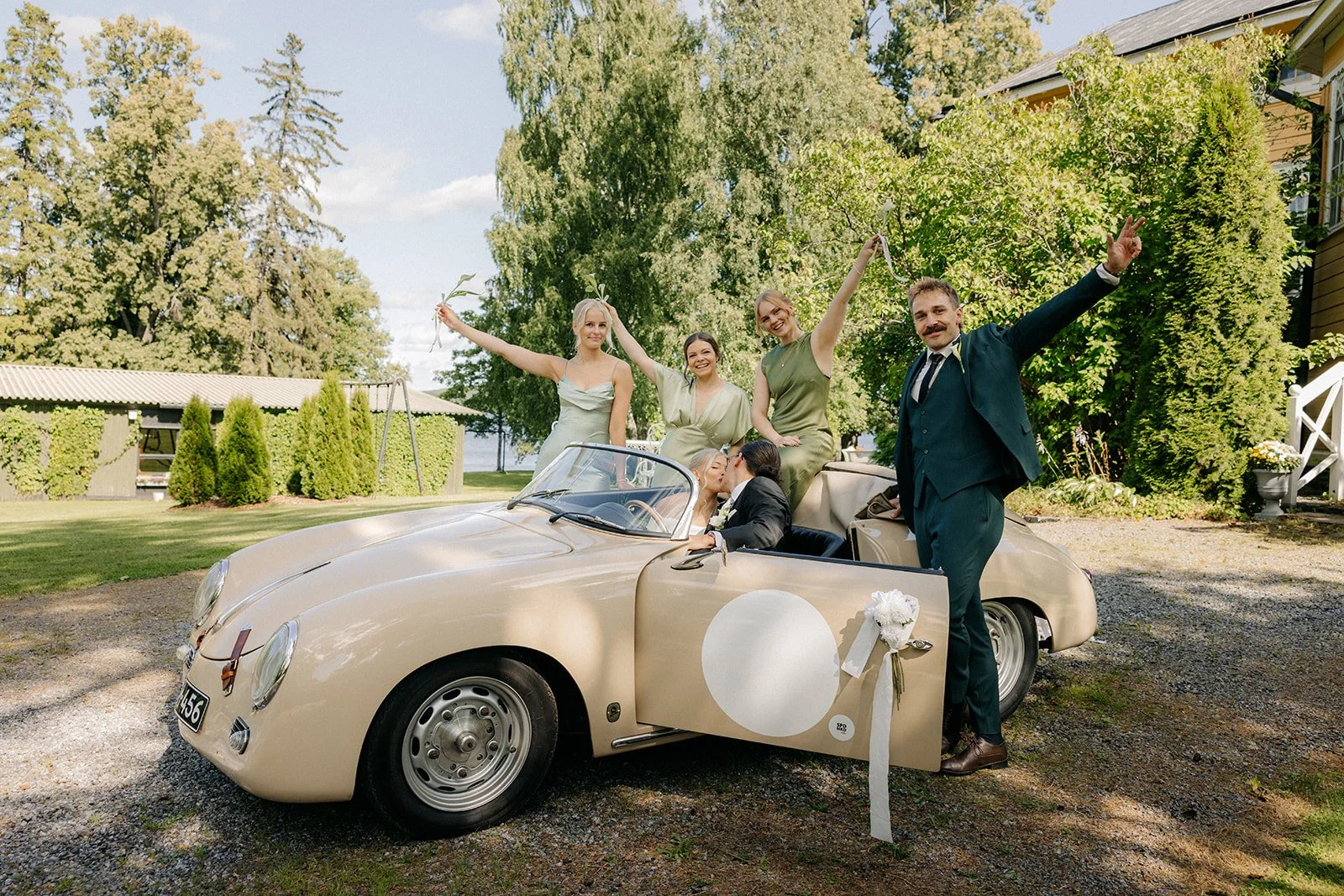 Iloiset kaasot ja bestman sekä suuteleva hääpari beigessä hääautossa.
