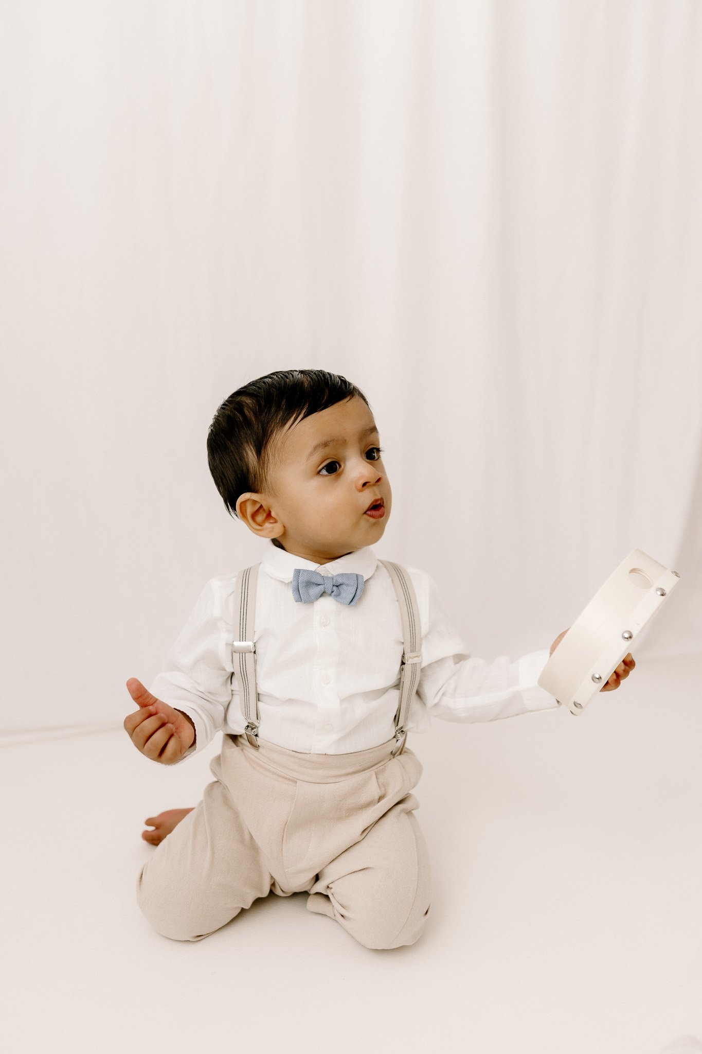 Young boy dressed in white shirt, beige pants with suspenders, and a blue bow tie, kneeling on the floor, holding a tambourine, with a white curtain backdrop.