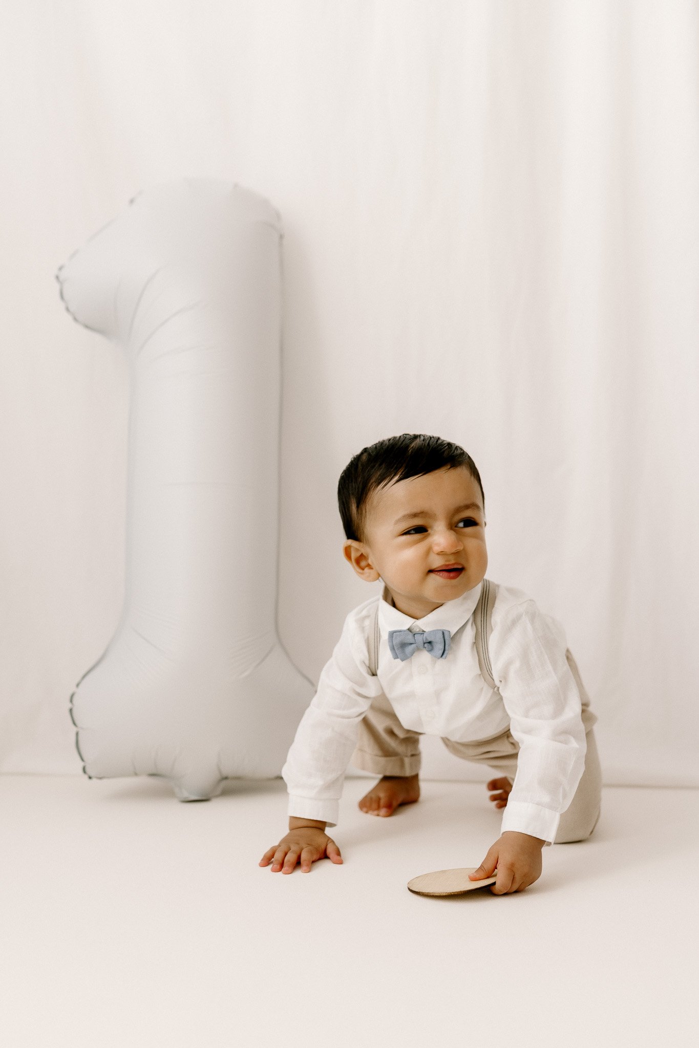A young child crawling on a white floor wearing a white shirt, beige pants, and a light blue bow tie, holding a small wooden object, with a large white number one balloon in the background.