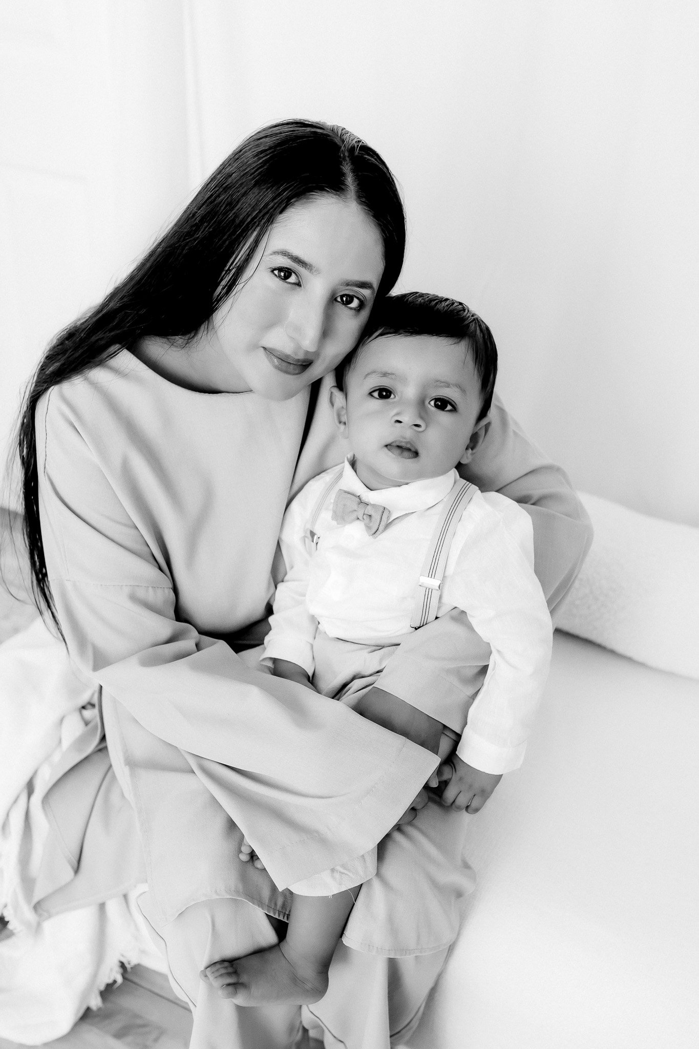 Black and white photo of a woman with long dark hair holding a young boy, both looking at the camera, sitting on a bed in a minimalist room.