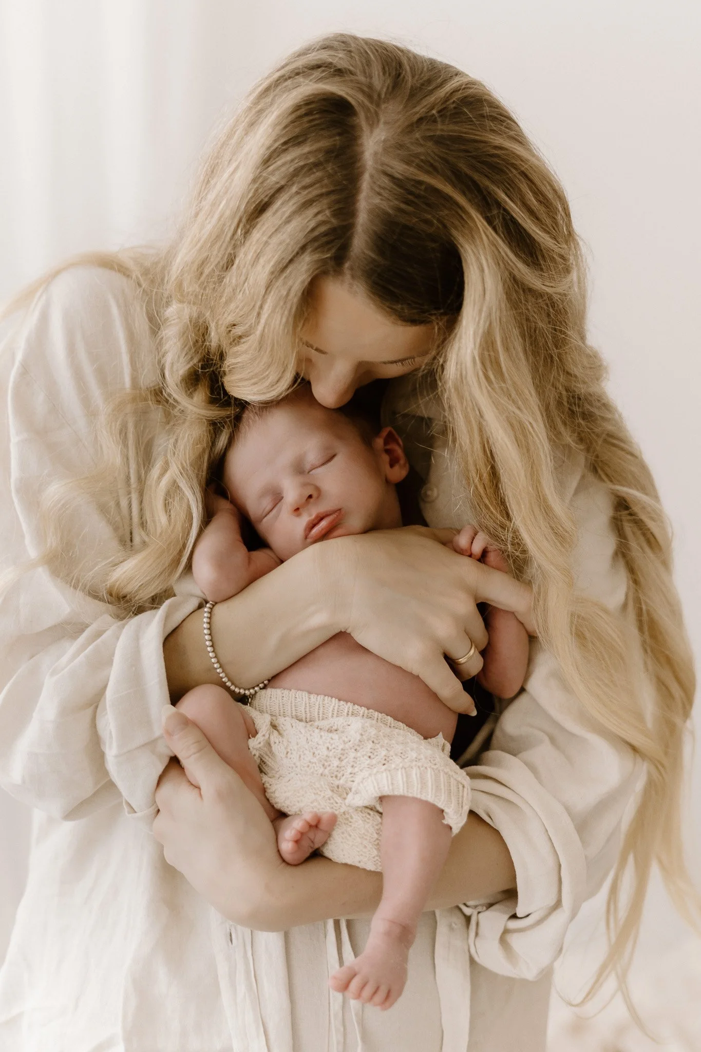 A woman with long blond hair holding a sleeping baby close to her chest, in a warm embrace.