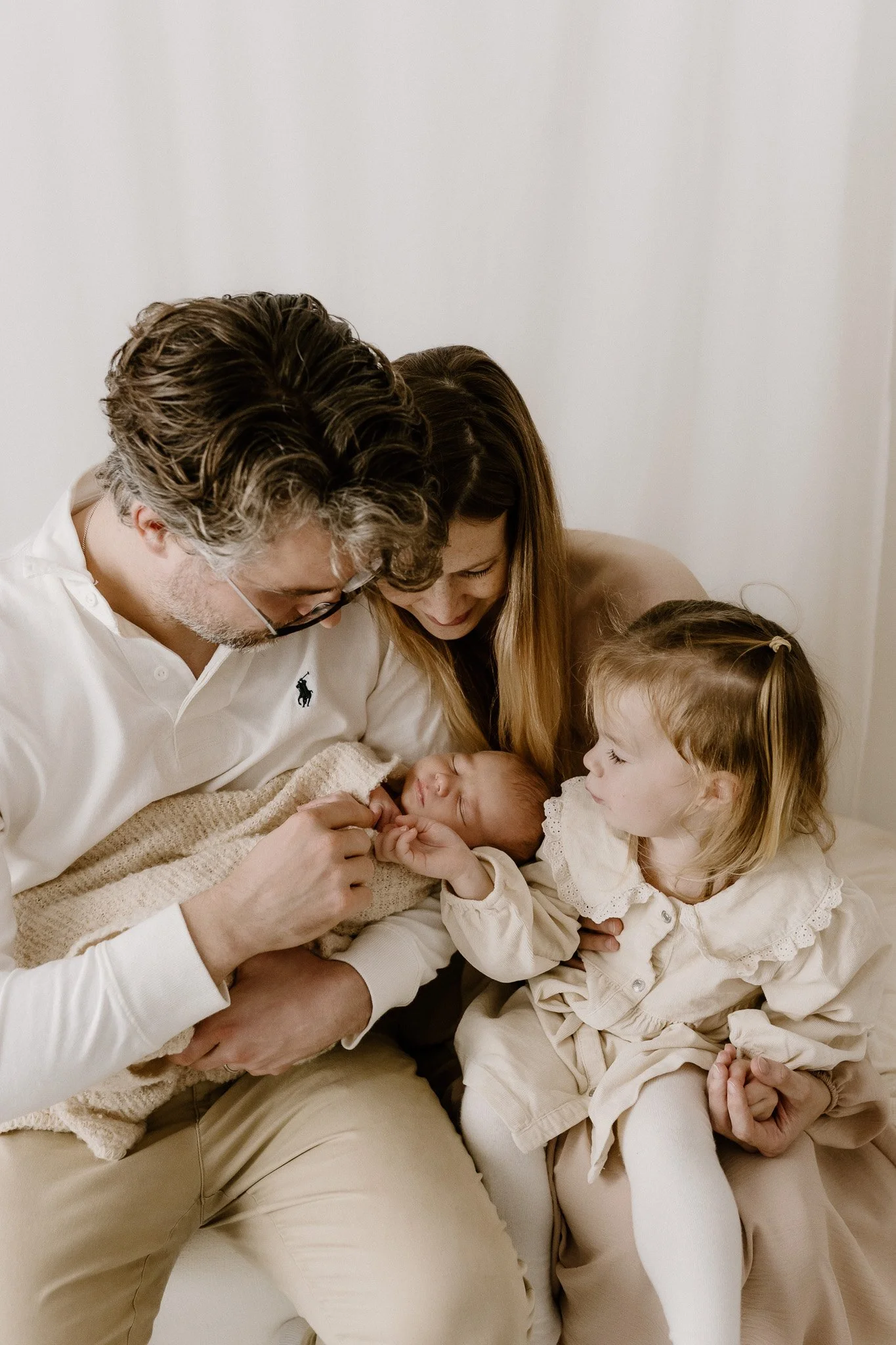 Family of four sitting together on a beige sofa in a cozy room, warmly gazing at their newborn baby who is wrapped in a beige blanket. The parents, a man with glasses in a white polo shirt and a woman with long hair in a neutral top, are gently holdi