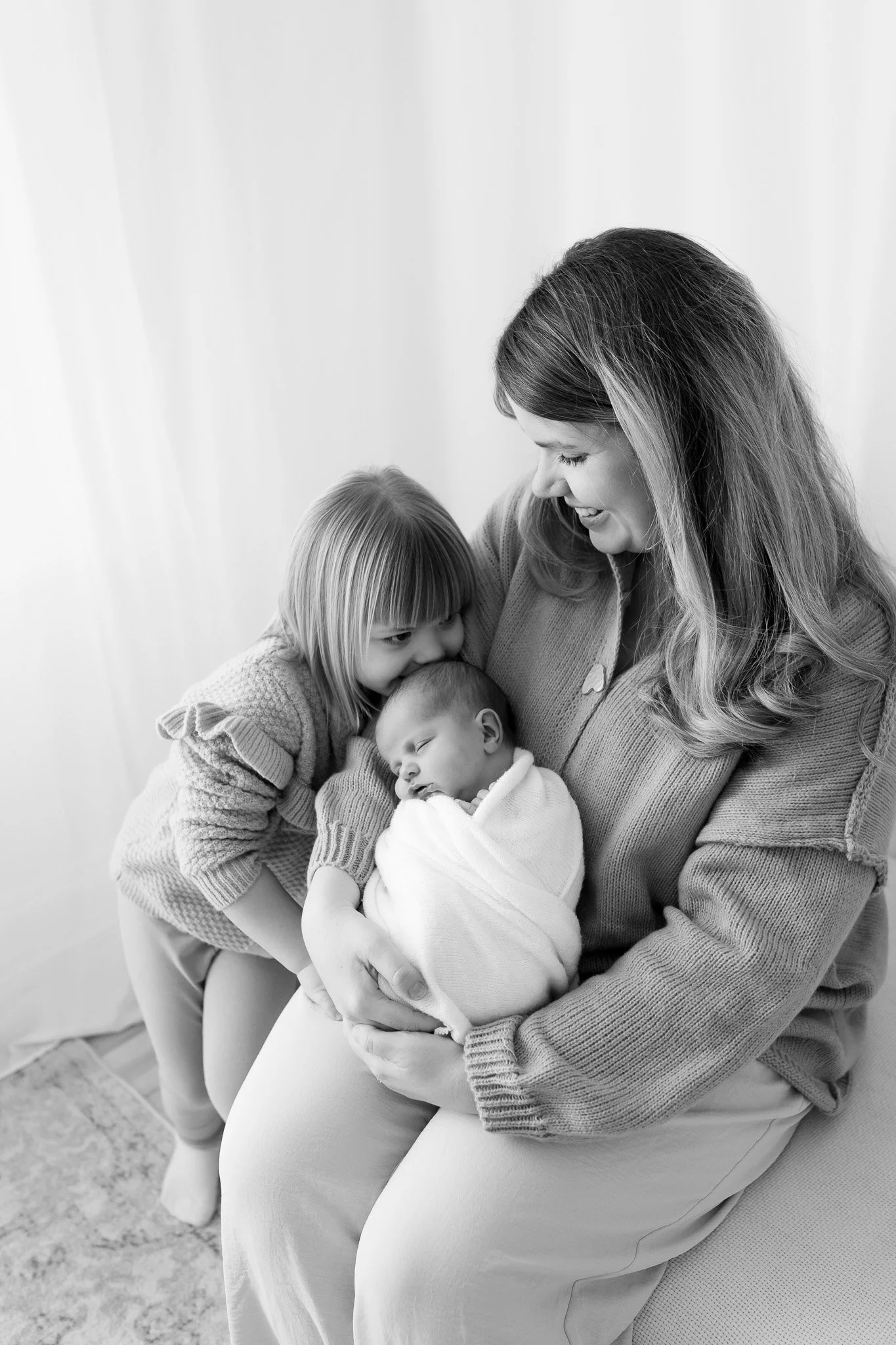 A woman holding a newborn baby wrapped in a blanket, with a young girl leaning in affectionately, all smiling.