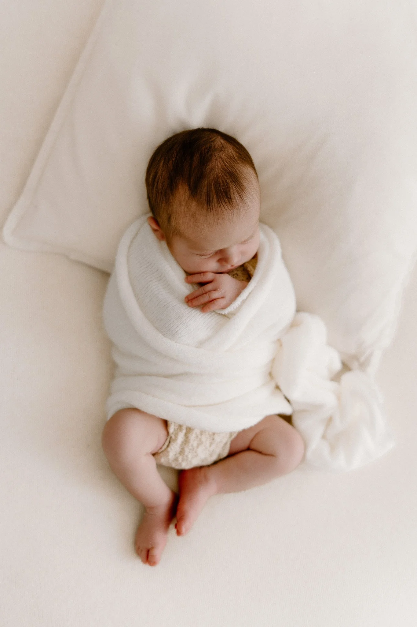 A sleeping baby wrapped in a white blanket, lying on a white bed with a white pillow.