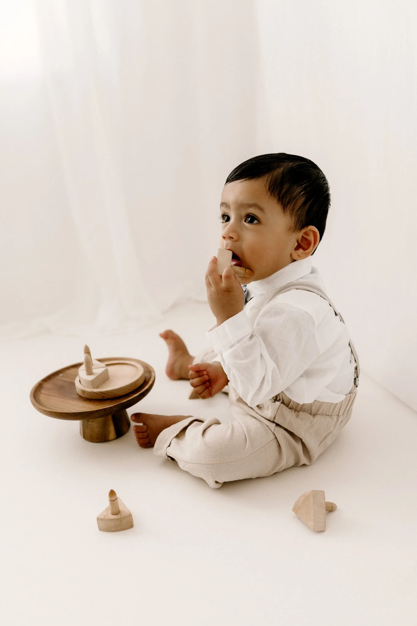 A young boy sitting on a white surface, playing with wooden toys, wearing a white shirt and beige pants.