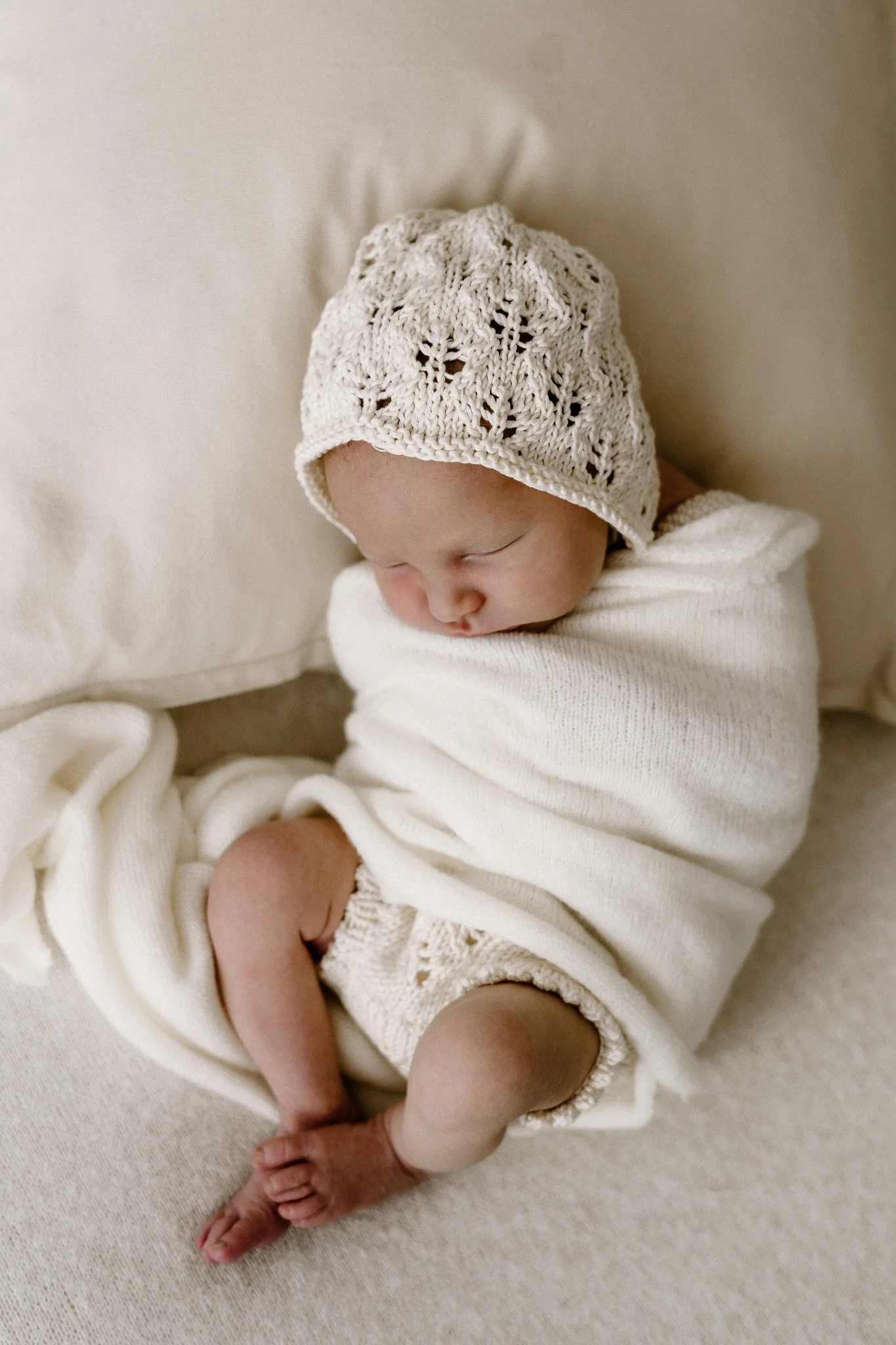 A newborn baby sleeping on a bed, wrapped in a white cloth, wearing a crocheted hat and diaper.