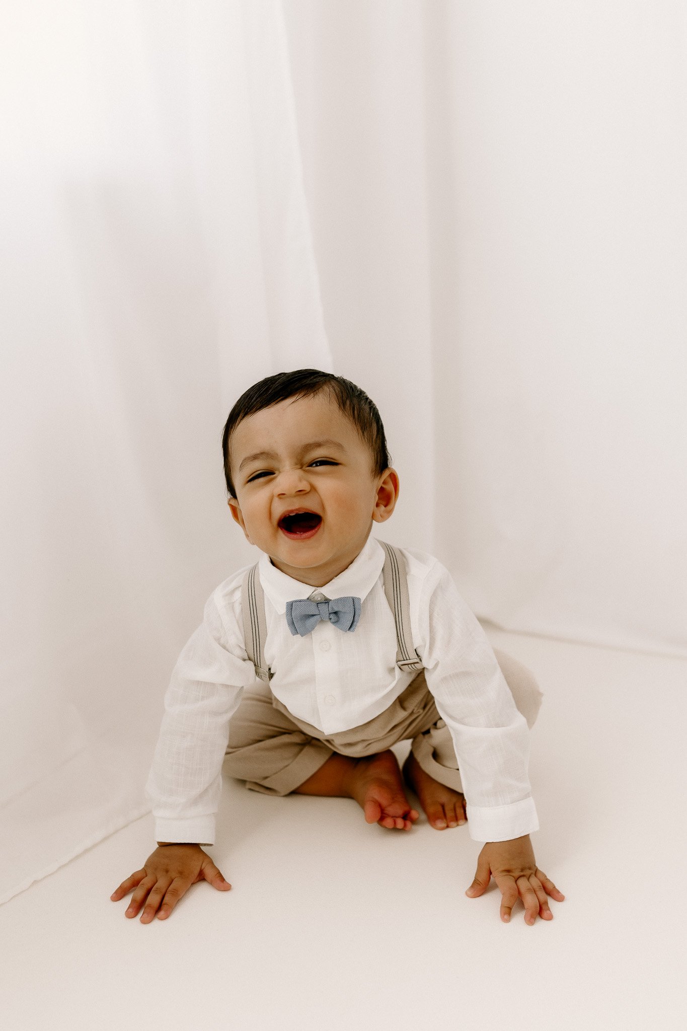A young boy in a white shirt with suspenders and a blue bowtie, sitting on the floor, smiling and laughing.