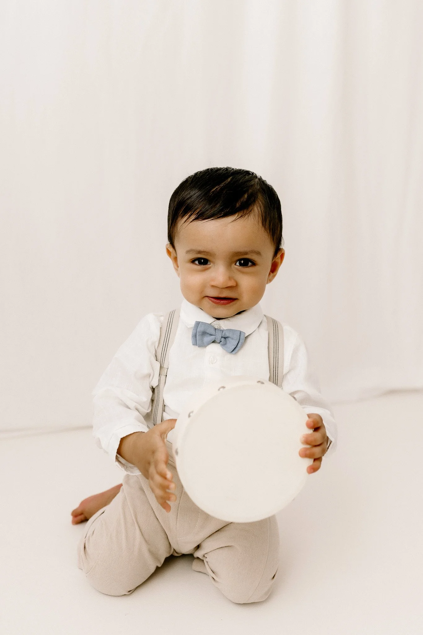 Young boy kneeling on the floor, holding a white drum, dressed in white shirt, beige pants, and a blue bow tie, with a neutral background.