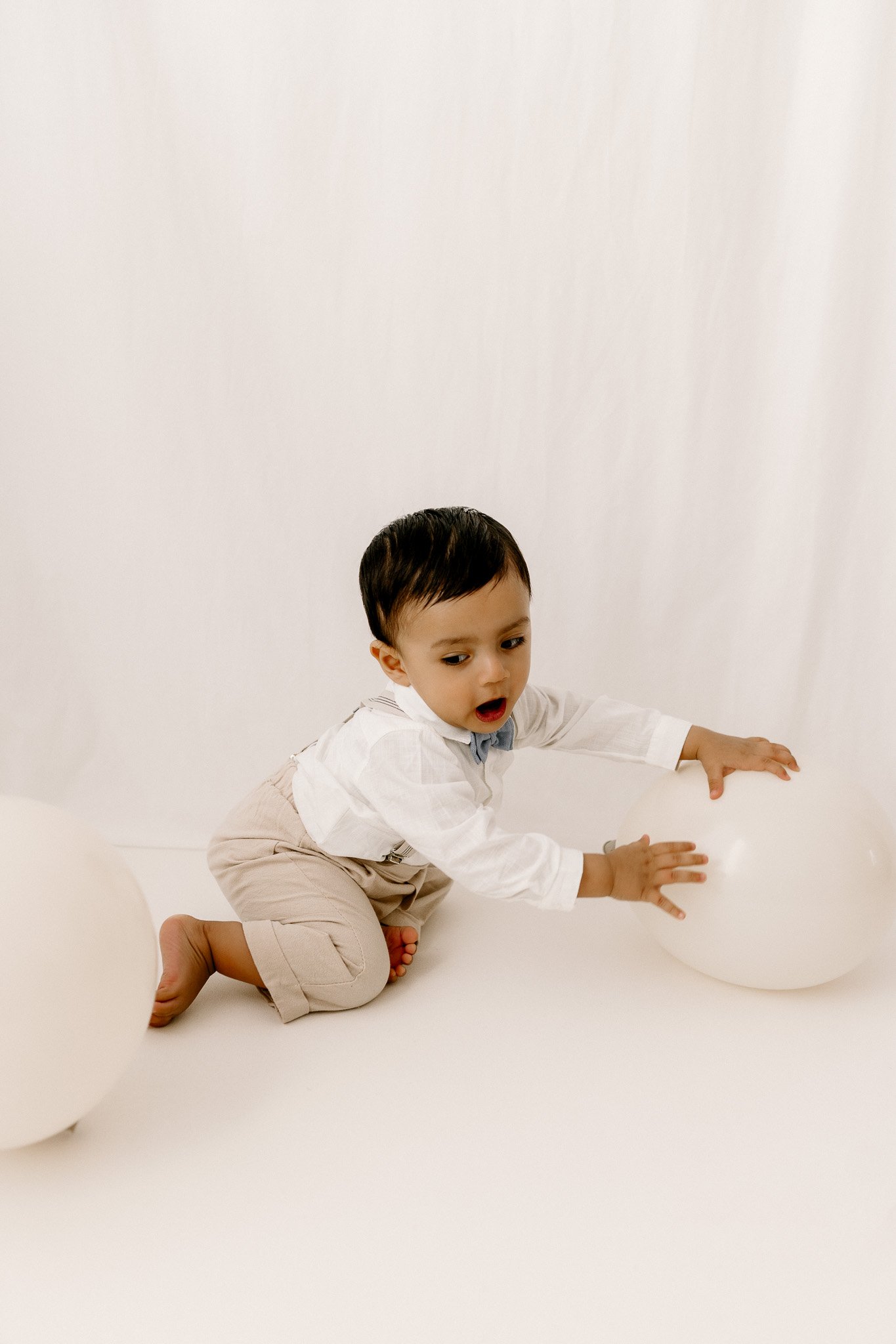A young boy with dark hair, dressed in a white shirt and beige pants, kneeling on a white surface and reaching for a white balloon in a blank white background.