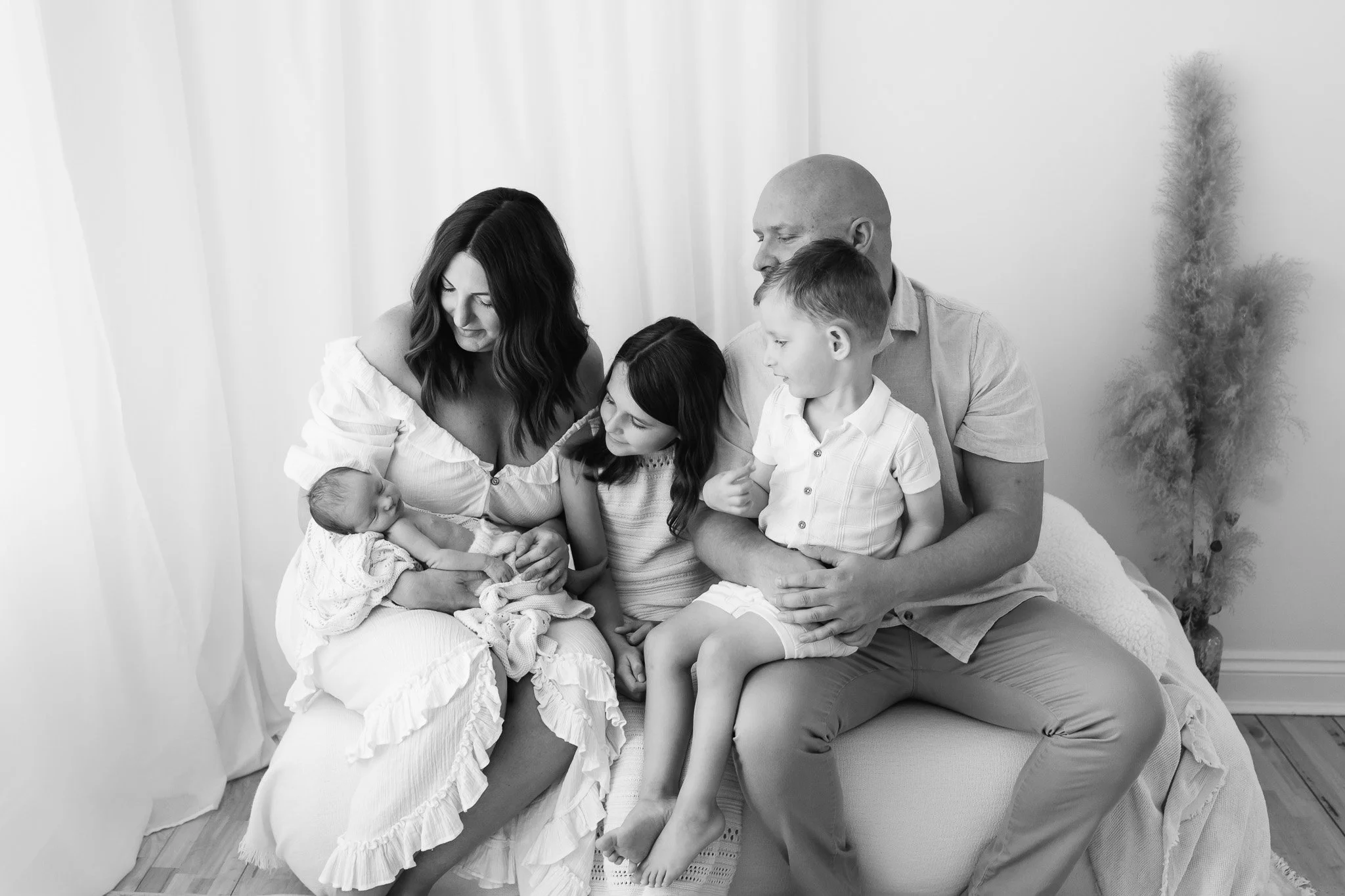 A family of five sitting on a bed, including a woman holding a newborn baby, a young girl, and a man with two young boys, in a bright room with a potted plant in the background.
