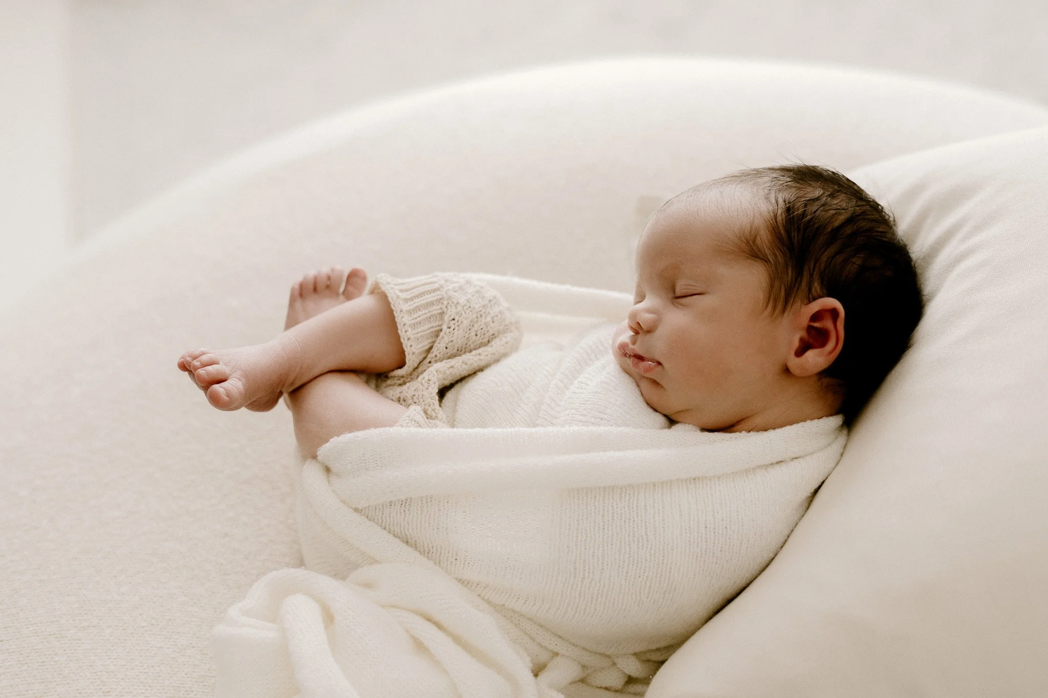 A newborn baby sleeping peacefully on a white pillow, wrapped in a soft white blanket and dressed in a knitted cream-colored outfit.