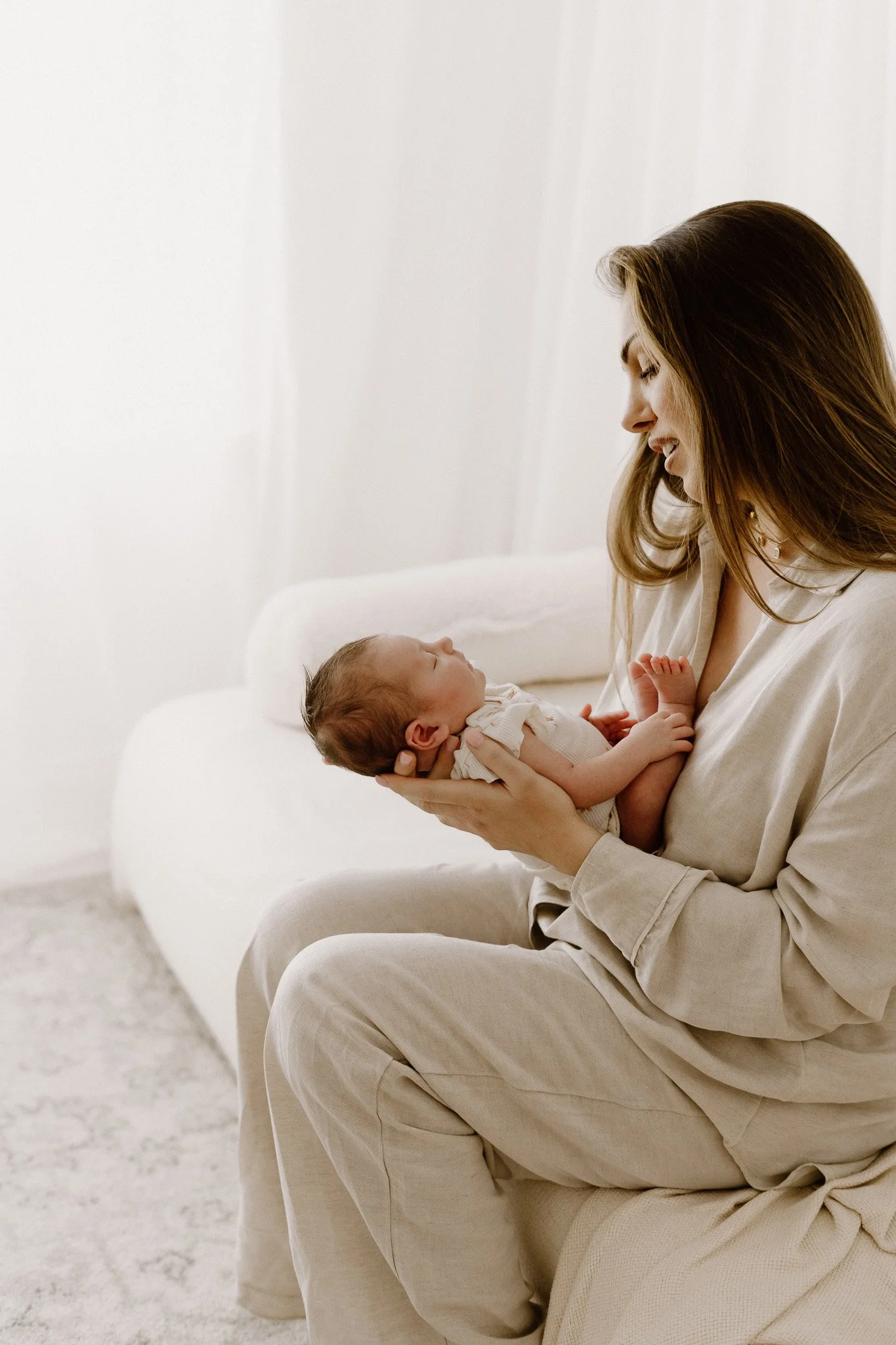 A woman holding a sleeping baby in her hands, sitting on a sofa in a neutral-colored room.
