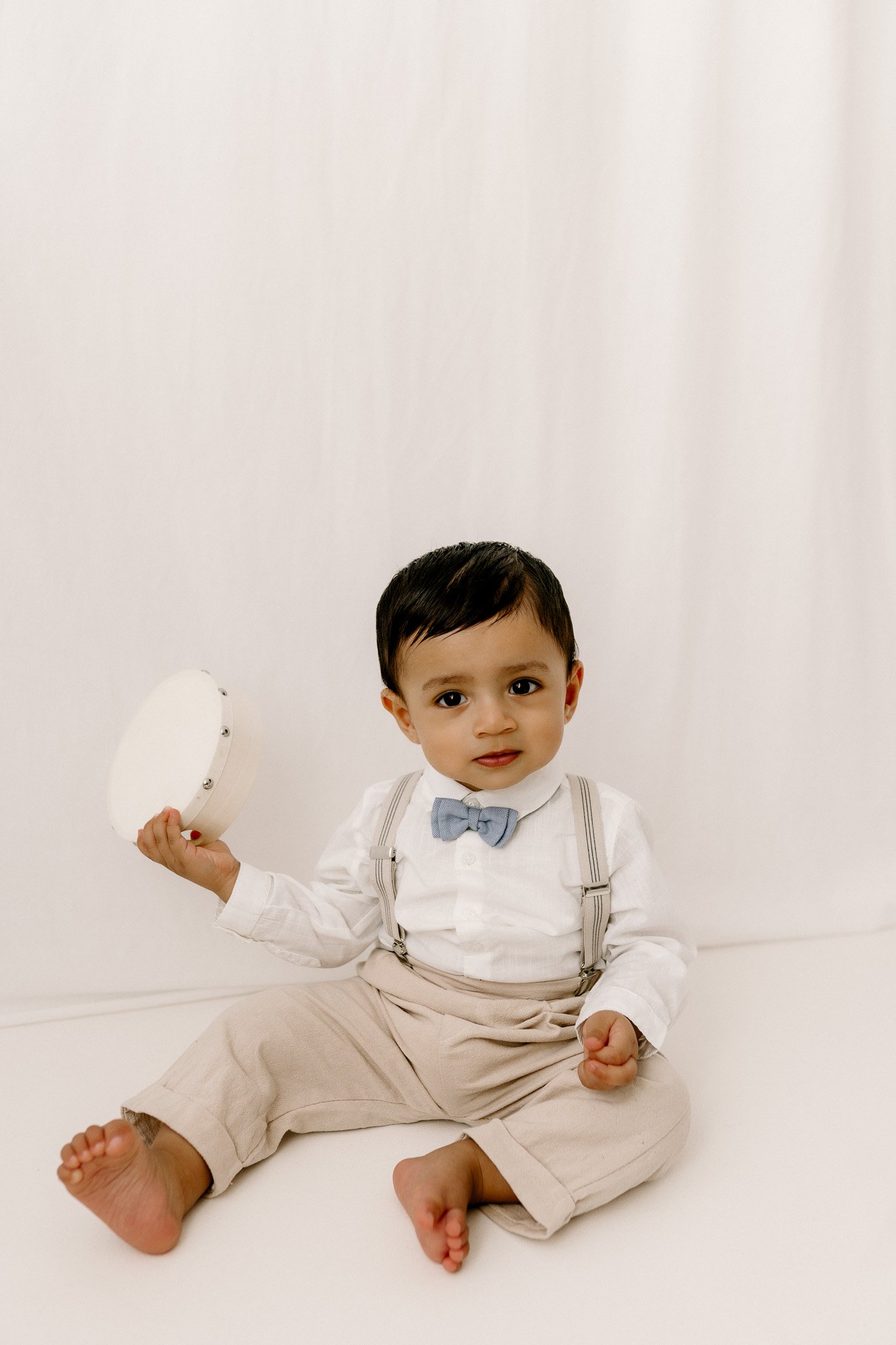 A young boy sitting barefoot against a plain white background, holding a small tambourine, wearing a white shirt, beige suspenders and pants, and a gray bow tie.