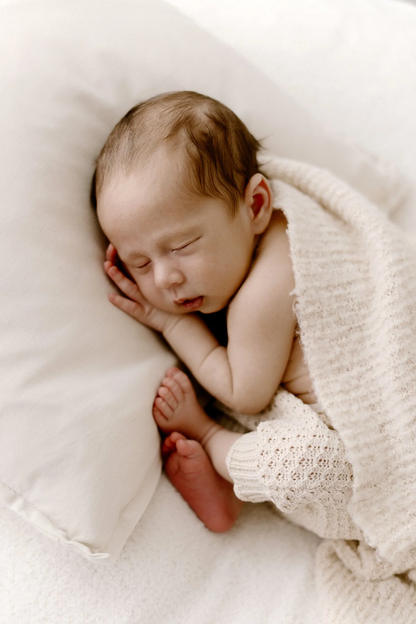 A sleeping newborn baby with light skin and brown hair, resting on a soft surface, partially covered by a knitted cream-colored blanket.
