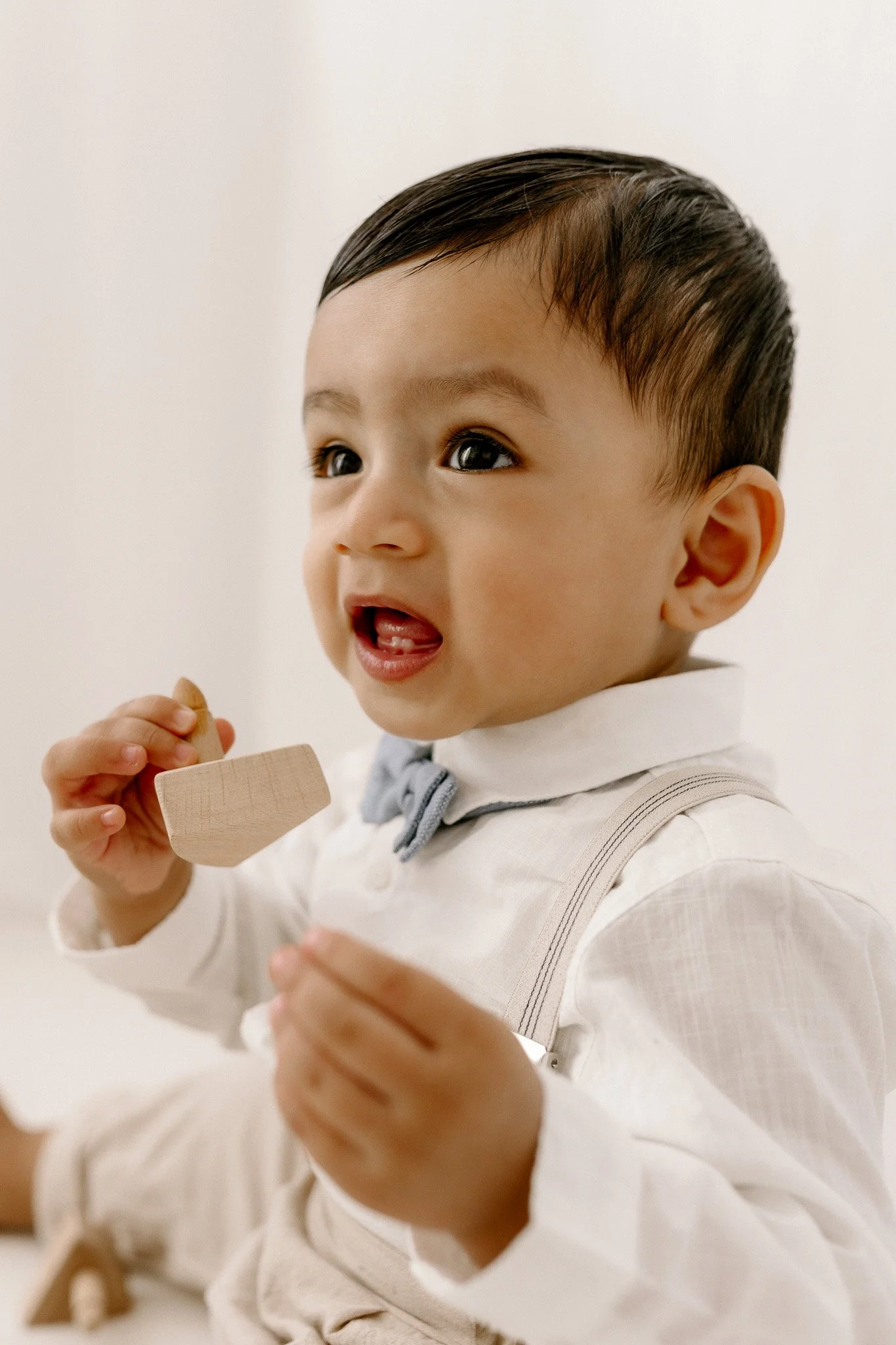 Close-up of a young Asian boy with dark hair, wearing a white shirt with suspenders and a bow tie, holding a small wooden toy and looking to the side.
